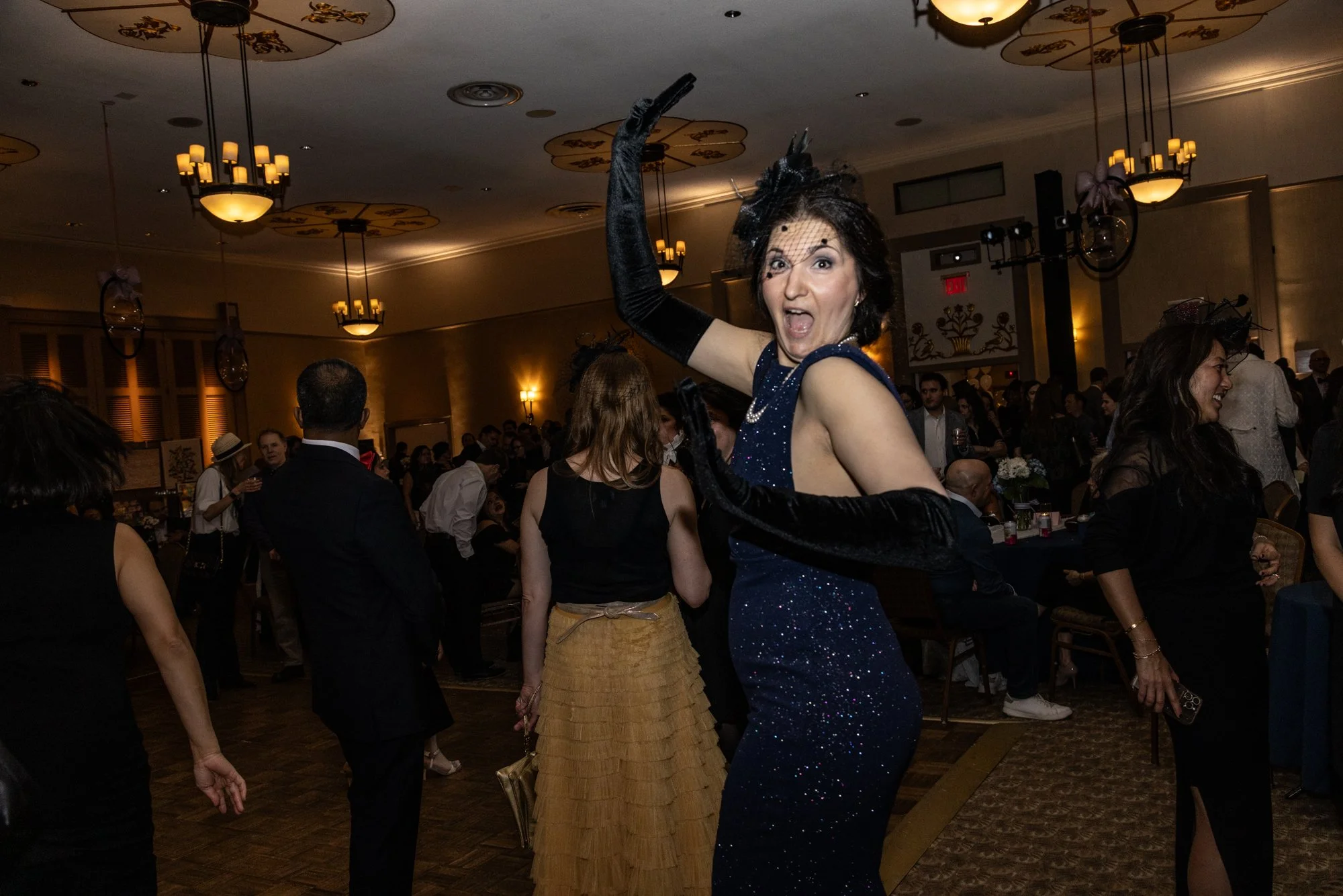 A woman in a dark blue, sparkling dress and black gloves dancing at a crowded indoor event, with guests and elegant decor in the background.