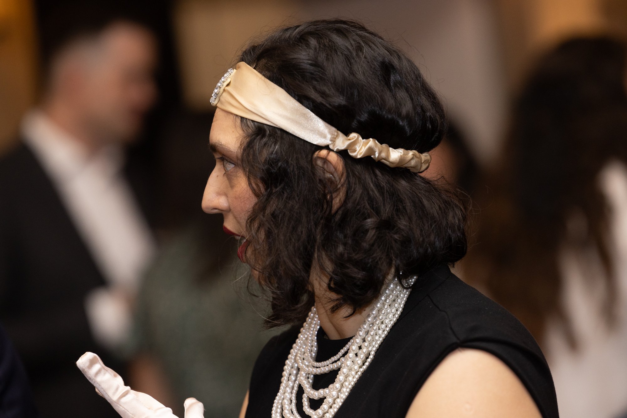 A woman with dark, curly hair wearing a satin headband with rhinestones, pearl necklaces, and a black dress participating in a formal event.