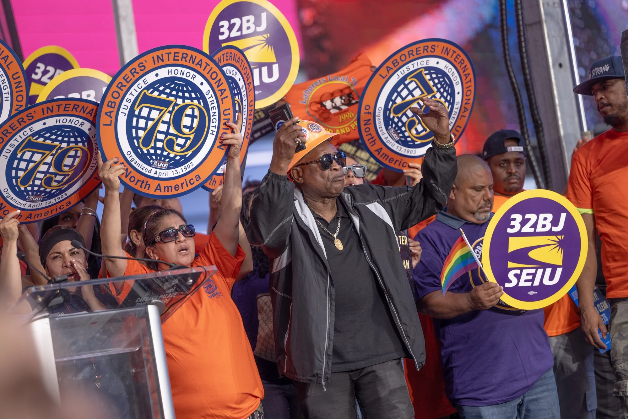 A group of people at a rally holding signs with the logo of SEIU, a labor union, and the number 79. The people are wearing orange and purple shirts, and some are wearing umbrellas and sunglasses.