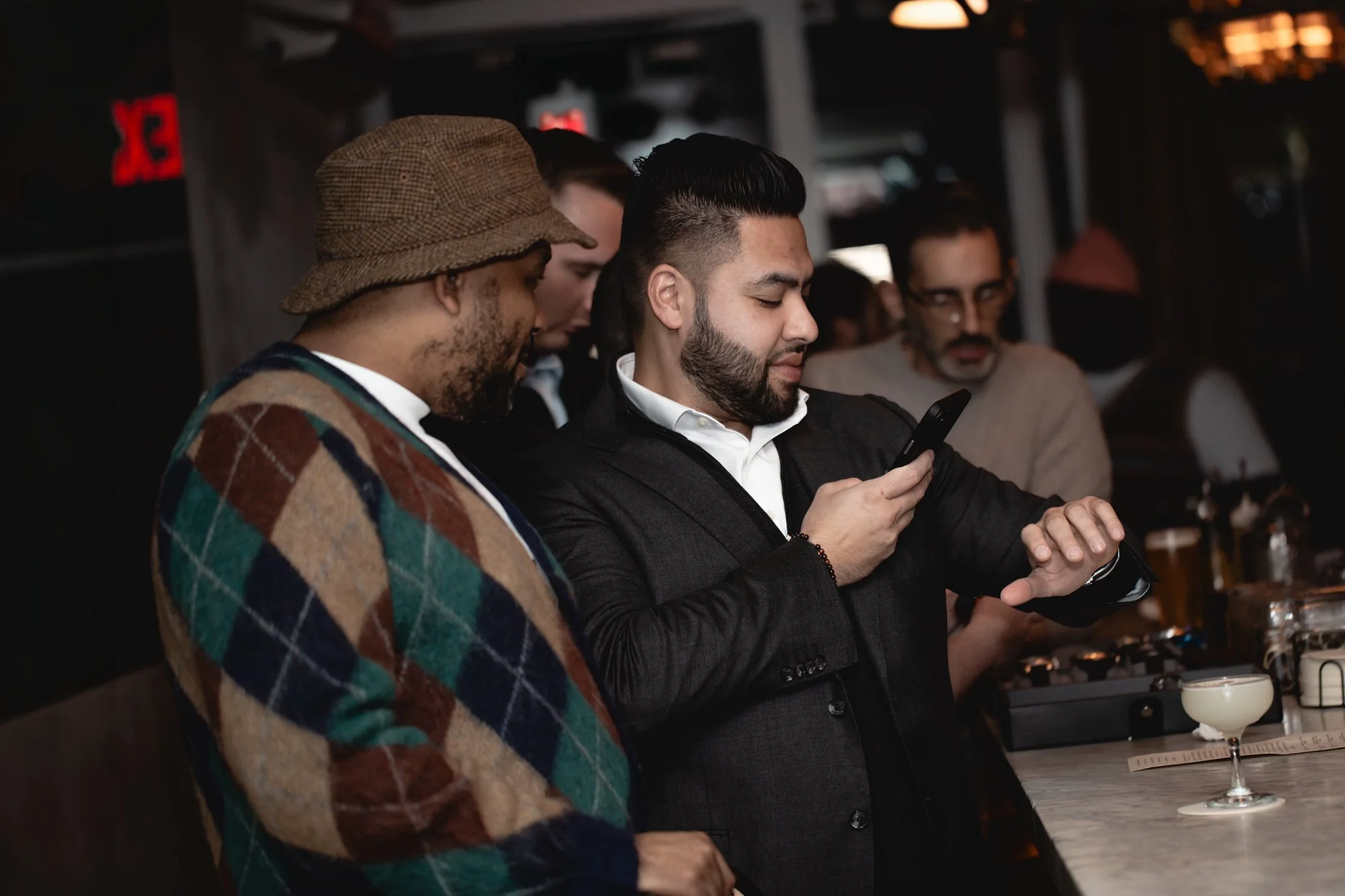 A man with black hair and a beard, wearing a dark blazer and white shirt, looking at his phone in a social setting. Next to him is another man wearing a brown plaid hat and a multicolored plaid jacket, also engaged with the phone. In the background, 