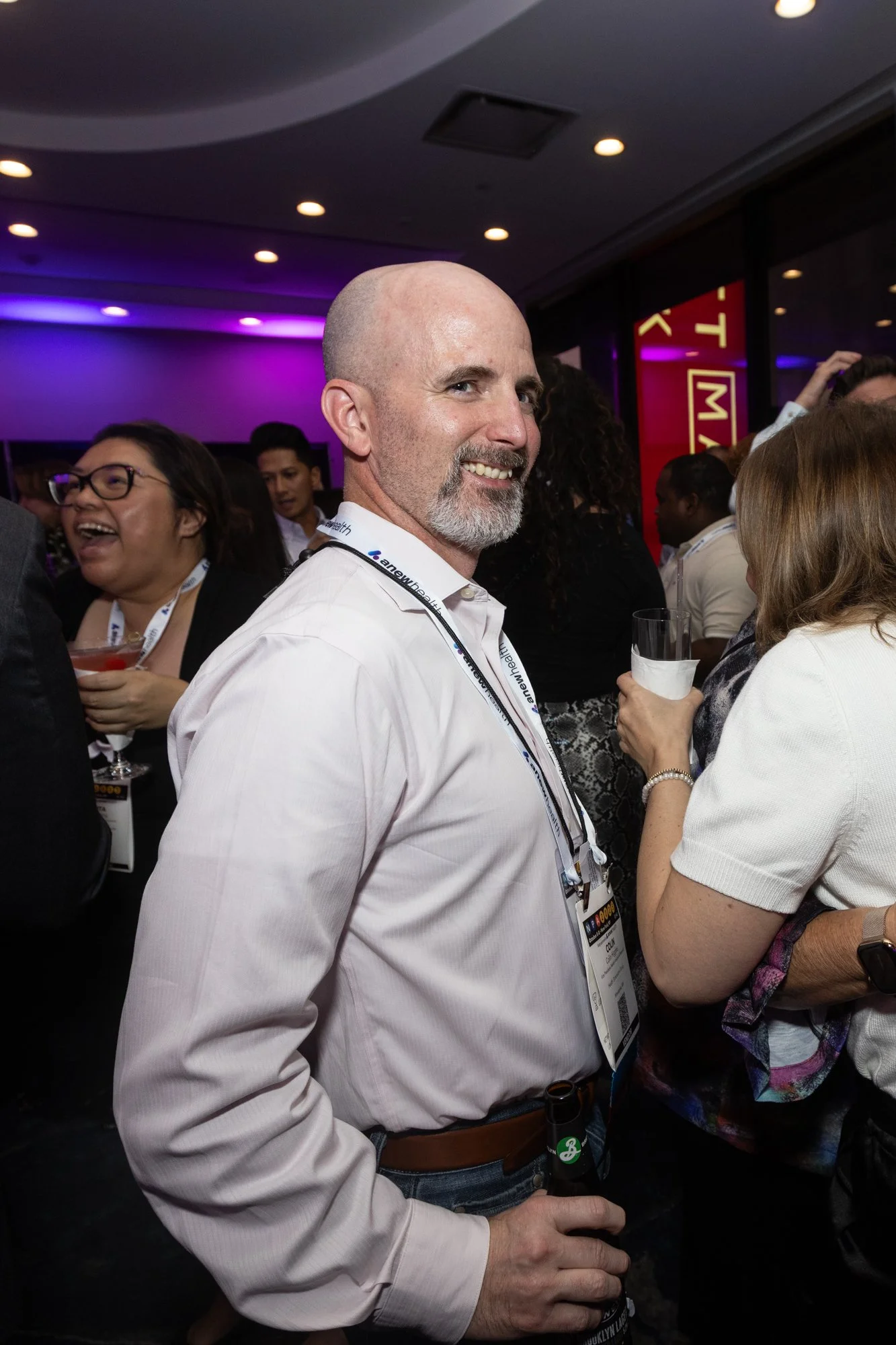A group of people at a crowded event, with a man in the foreground smiling and holding a beer, wearing a white shirt and conference badge, amidst others engaging and laughing.