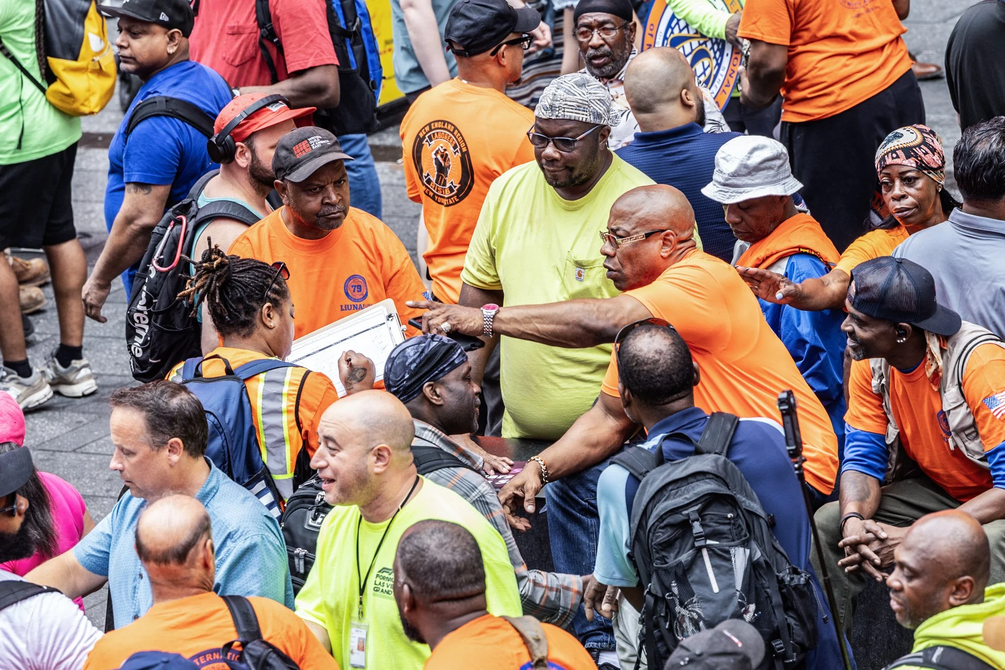 A crowd of people gathered in a street, many wearing orange shirts and some with backpacks. People are engaging in conversation, and there are various expressions of focus and interaction among them.