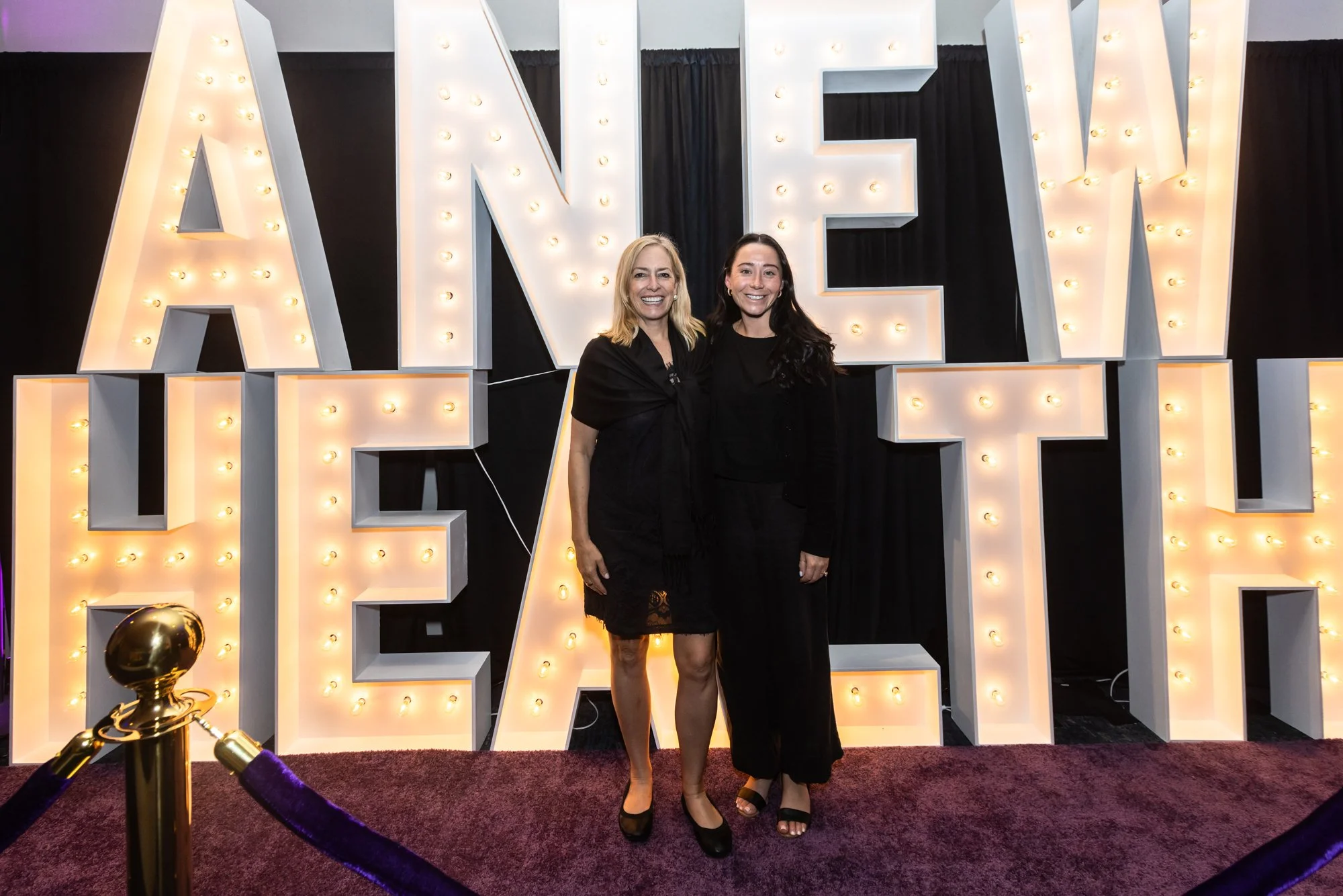 Two women in black dresses standing in front of a large illuminated sign that spells 'LOVE' at a formal event or celebration, with a velvet rope barrier and dark curtain backdrop.