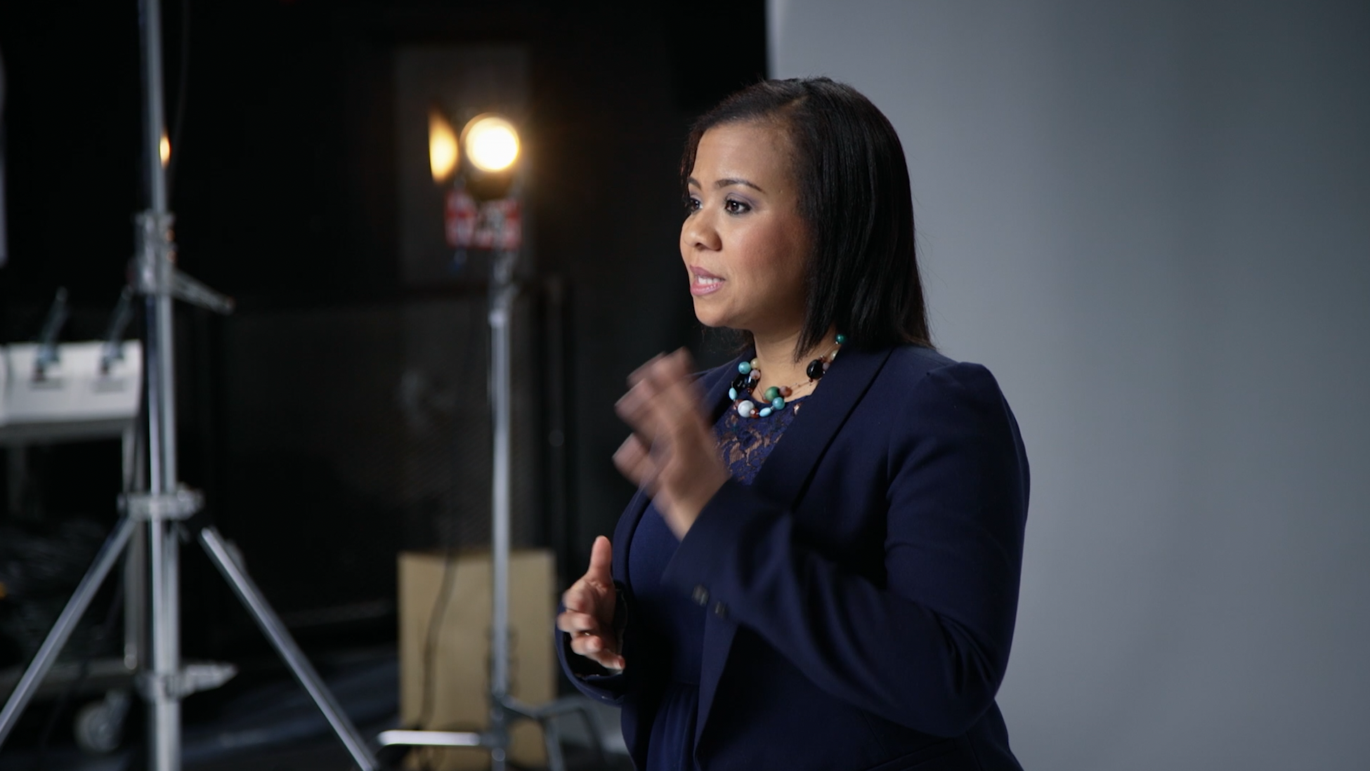 A woman with dark hair, wearing a dark blazer and a colorful beaded necklace, speaking or giving a presentation in a studio setting with lighting equipment visible in the background.