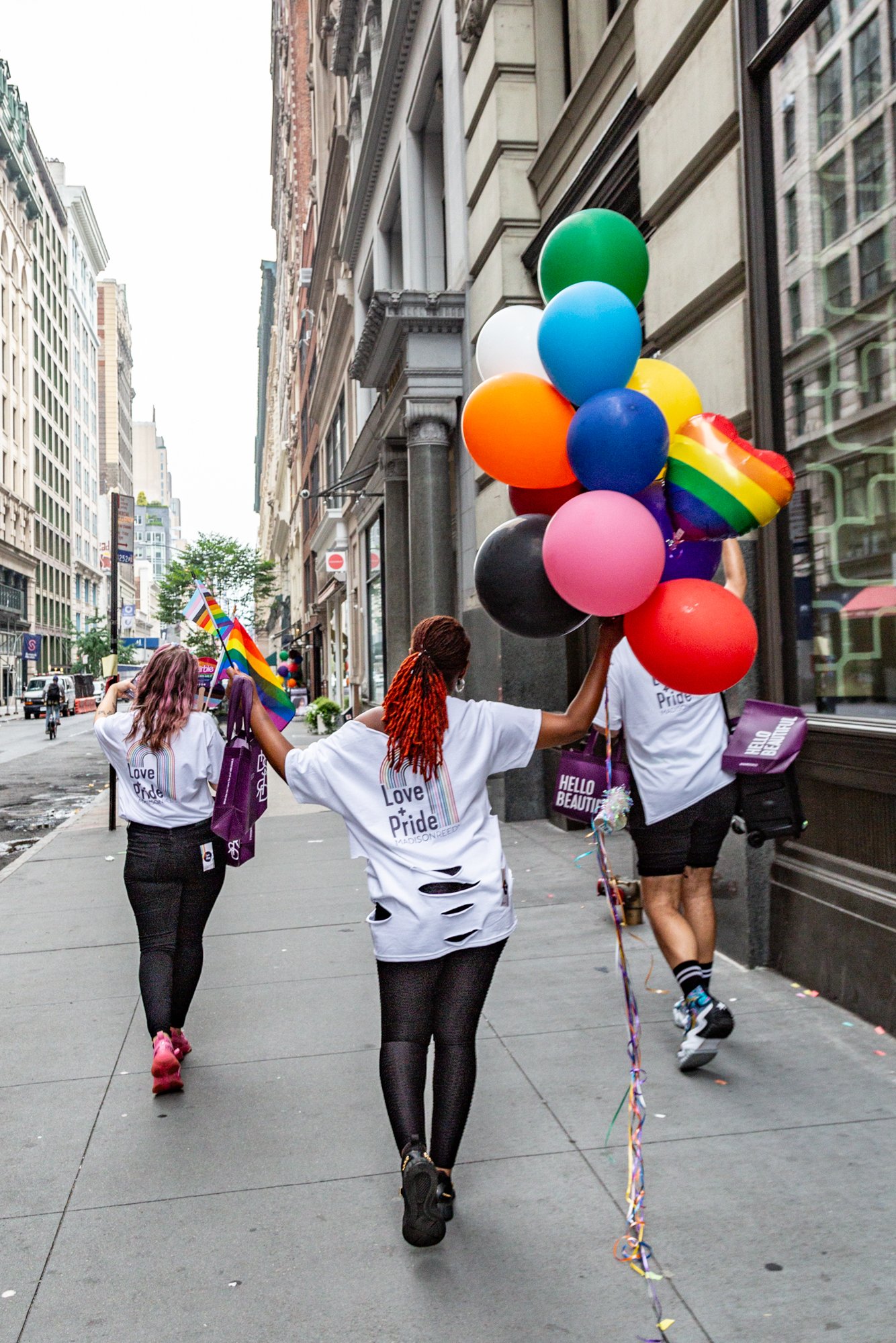 People celebrating Pride on a city sidewalk with rainbow flags, colorful balloons, and Pride shirts.