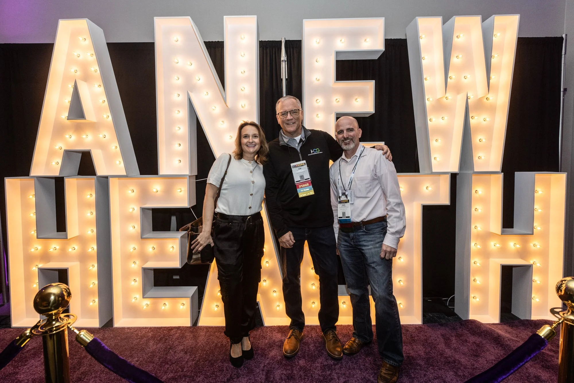 Three people, two men and one woman, smiling and standing together in front of large illuminated letters spelling 'LOVE.' The group is at an event, as indicated by their badges and the velvet rope stanchions in the foreground.