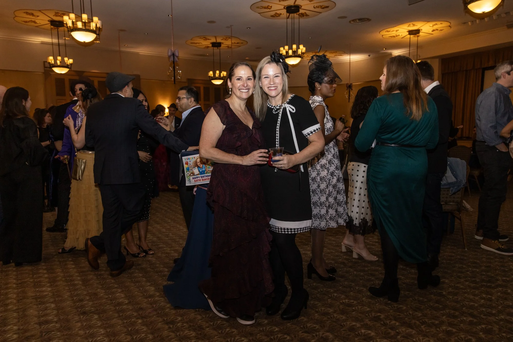 People at a social gathering in a ballroom, two women in the center smiling at the camera, holding drinks, dressed in party attire. Other guests socialize in background under warm lighting.