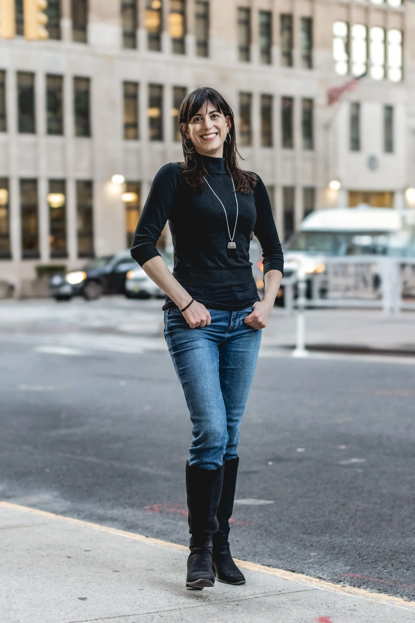 A woman with dark hair, wearing a black turtleneck sweater, blue jeans, and black boots, stands on a city street with a big smile, headphones around her neck, in front of a modern office building.