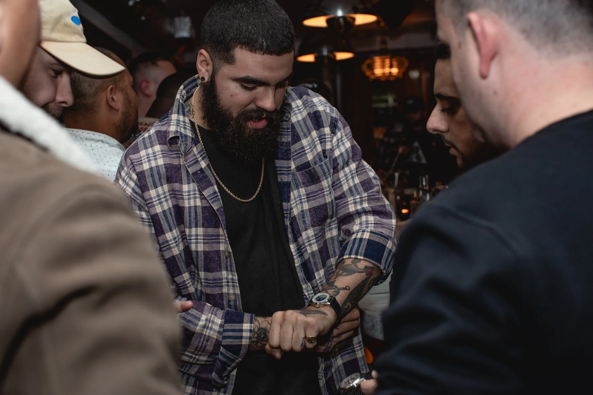 A group of people in a dimly lit room with four visible men. One with a beard and short hair, wearing a plaid shirt, black t shirt, gold chain, and tattoos on his left forearm, looks down with hands clasped. Others are partially visible, with one wea