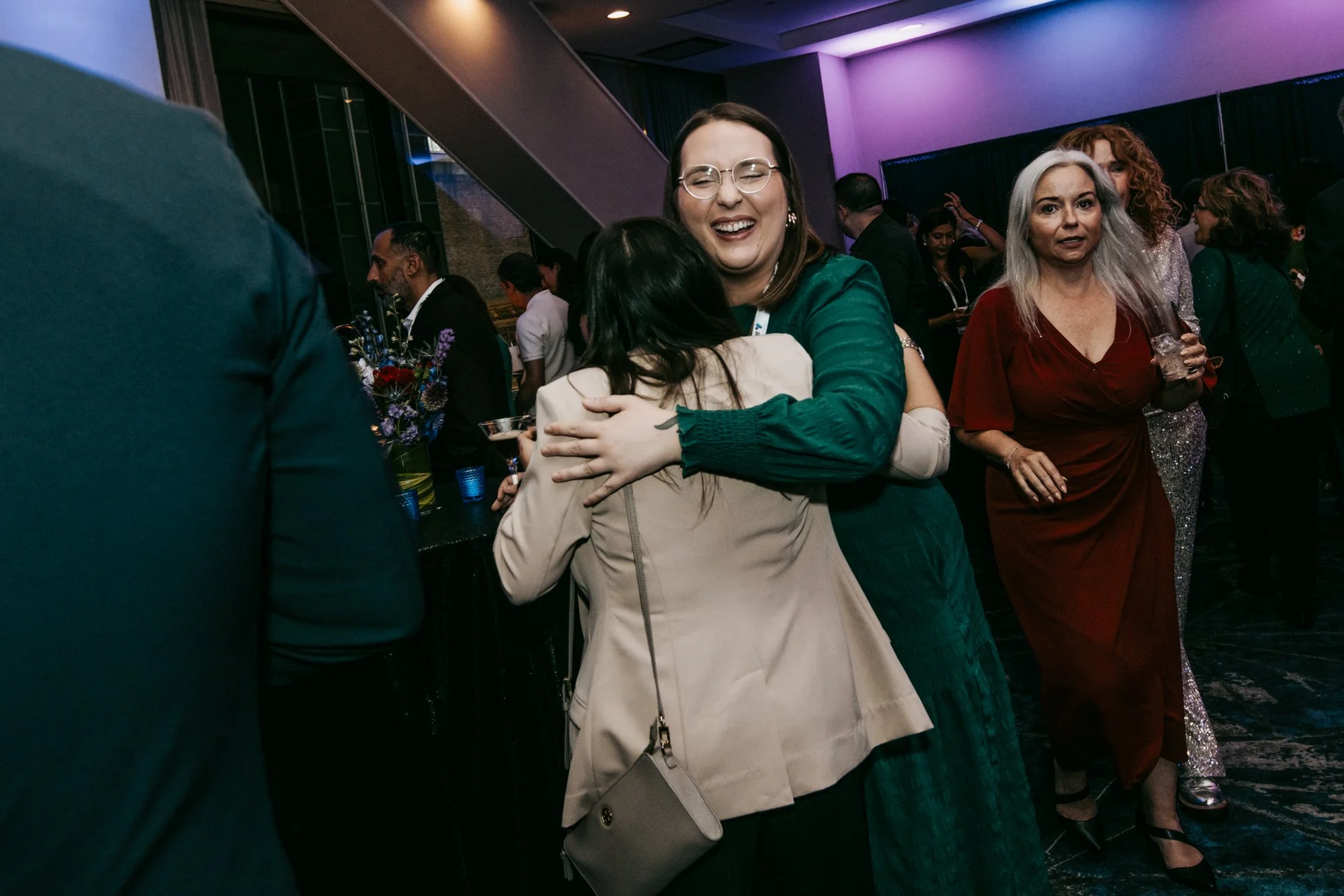 People at a networking event or conference, with one woman in a green dress hugging another person, others in the background socializing, with event lanyards and drinks.