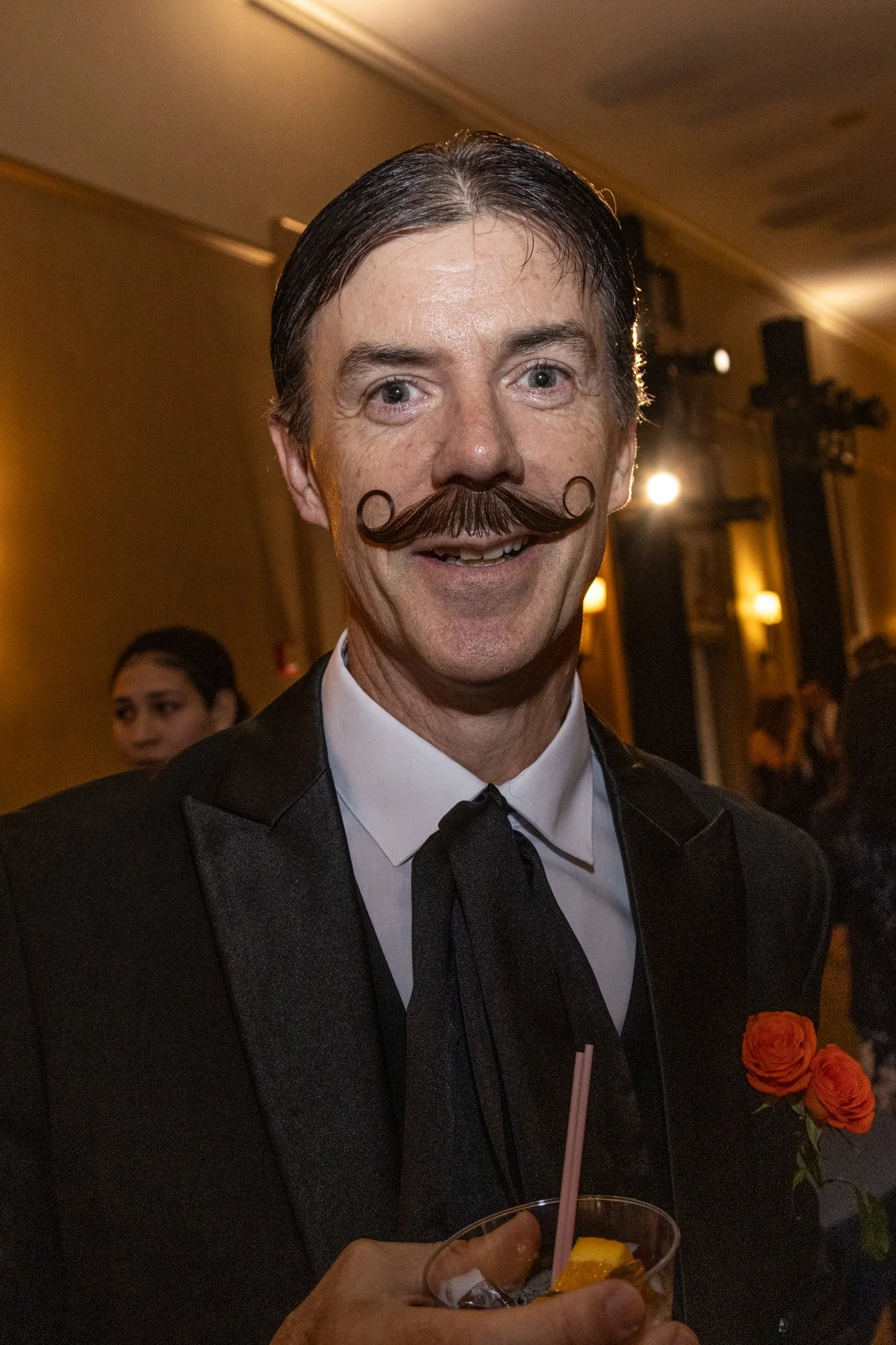 Man with a vintage styled mustache, dressed in a tuxedo at a formal event, holding a drink with fruit garnish, with a woman and orange roses visible in the background.
