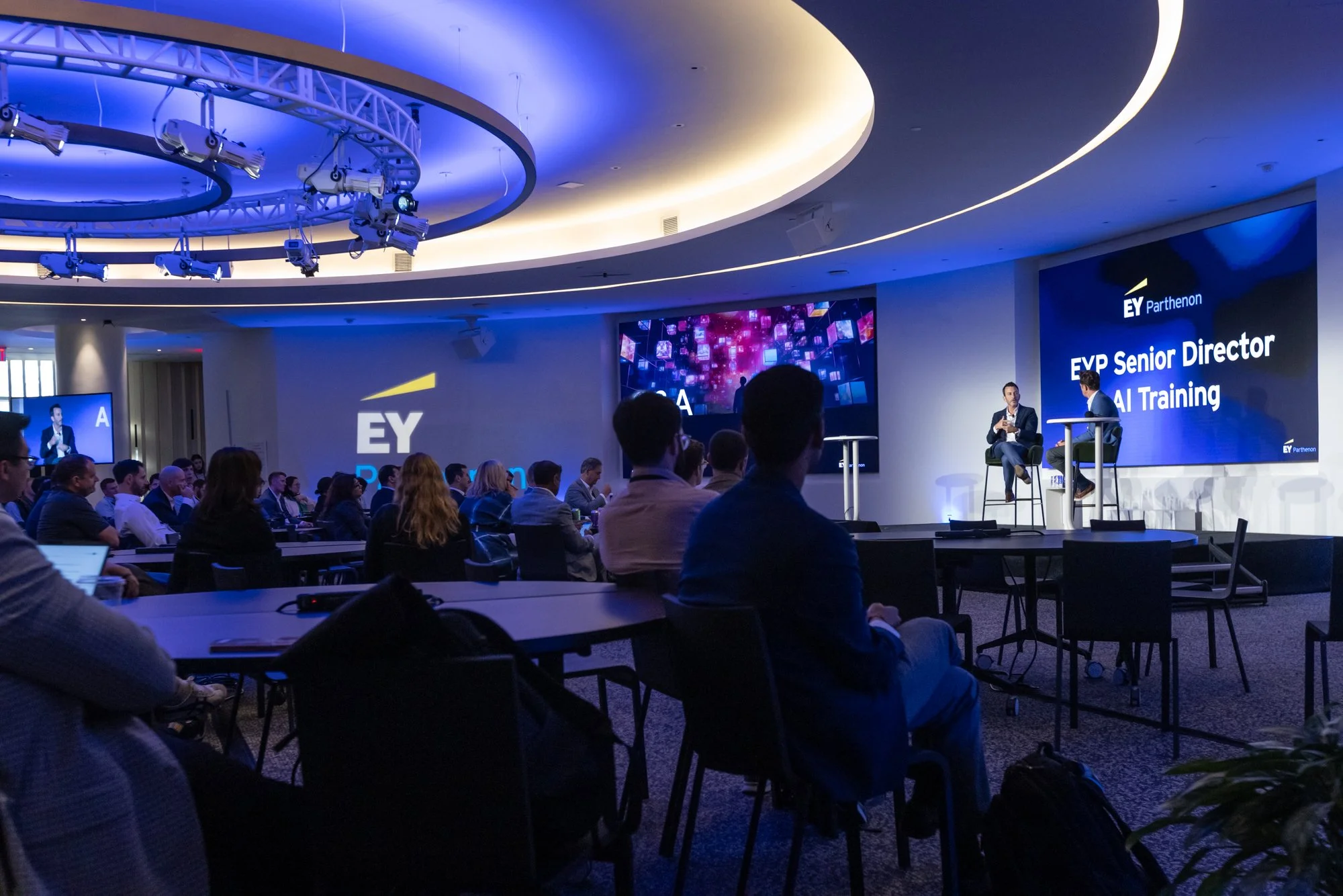 A large conference room filled with attendees sitting at tables, listening to speakers on stage. The stage has large screens displaying the EY logo and event details like 'EVP Senior Director AI Training.' The room is well-lit with modern ceiling lig