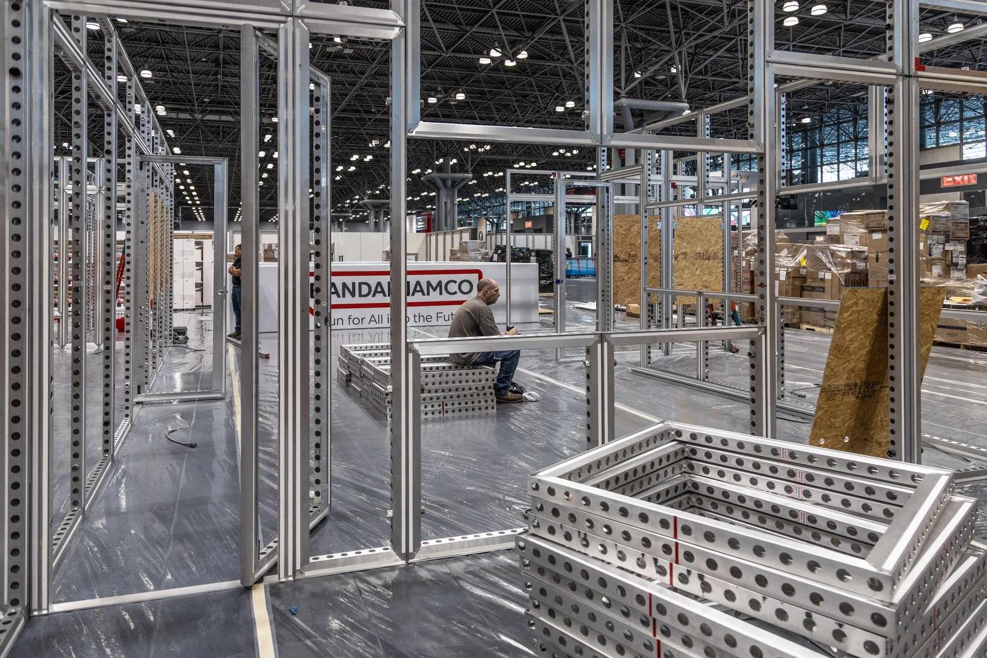 Workers assemble aluminum framing and display structures at a large trade show or convention center.