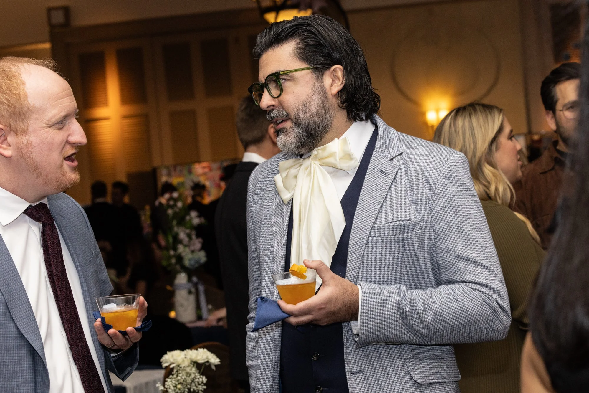 Two men in suits converse at a social event, each holding a cocktail glass garnished with an orange slice. The background shows other guests and a warmly lit room with floral arrangements.
