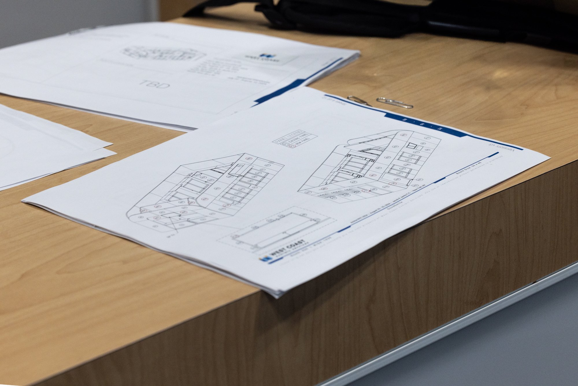 Architectural floor plans and technical drawings spread across a wooden desk with paper clips