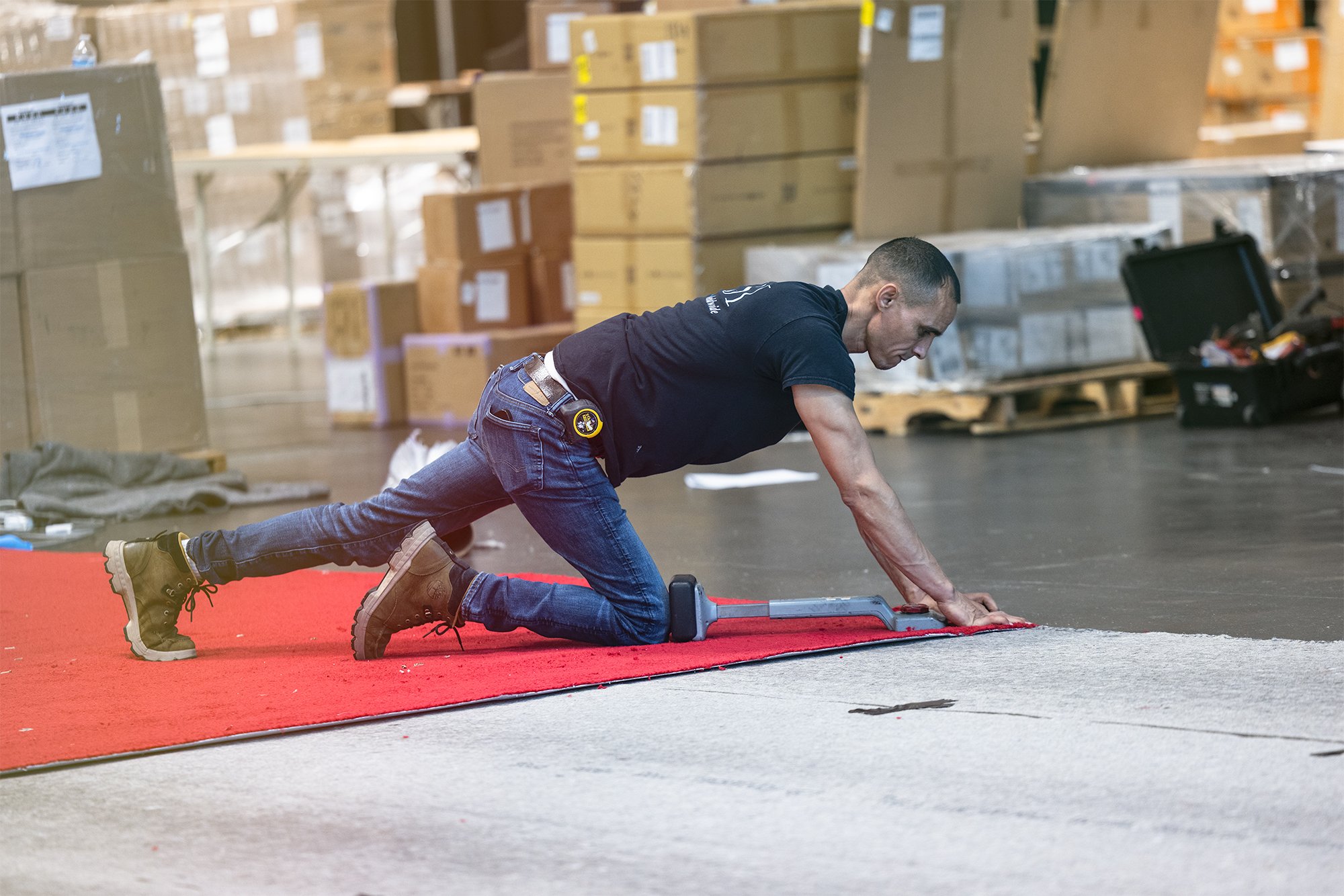 Man performing a stretching exercise on a red mat in a warehouse filled with cardboard boxes