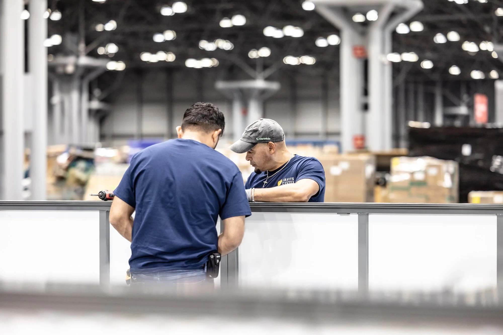 Two workers in navy shirts assembling equipment at a trade show booth inside a convention center.