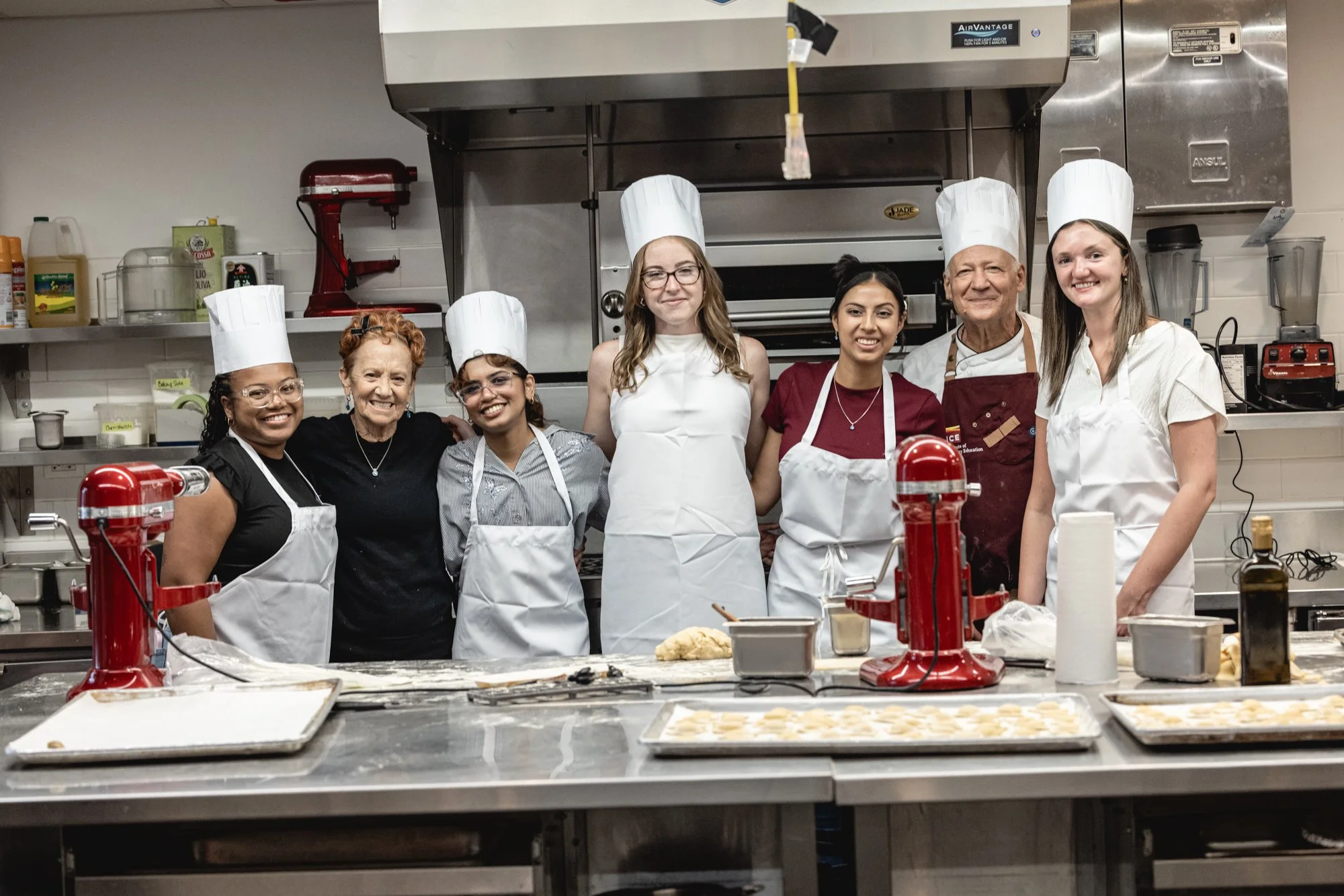 A diverse group of seven people, including both women and men, wearing white chef hats and aprons, standing together in a commercial kitchen. They are smiling and appear to be celebrating or posing after a cooking or baking activity. In the foreground, there are trays with baked goods and the kitchen is equipped with various appliances and ingredients on shelves.