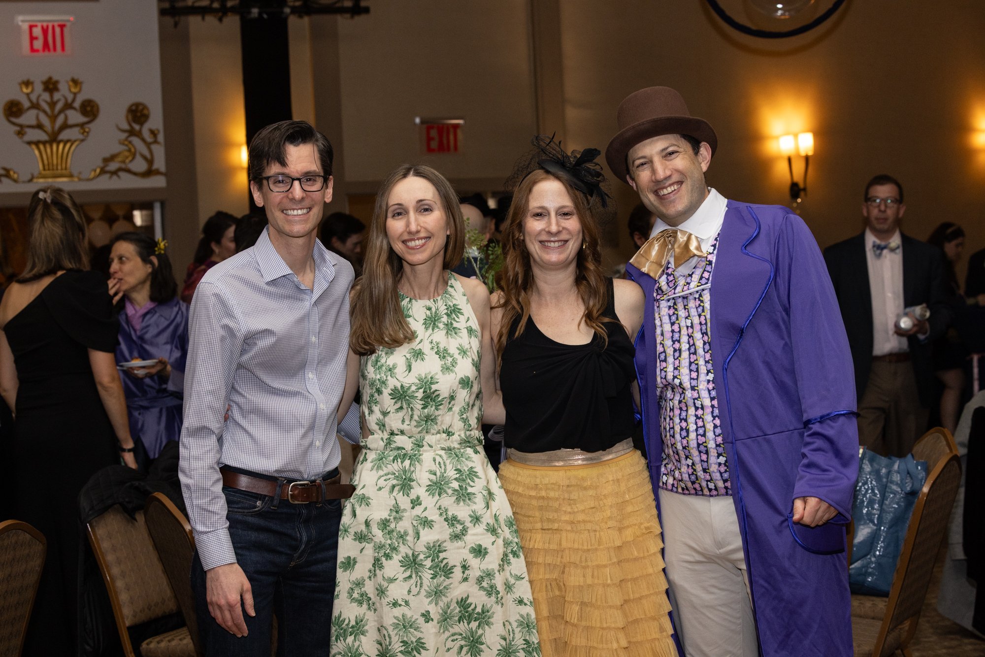 Four people standing together at an indoor event, smiling at the camera; two men and two women, dressed casually and one in a colorful costume with a hat.