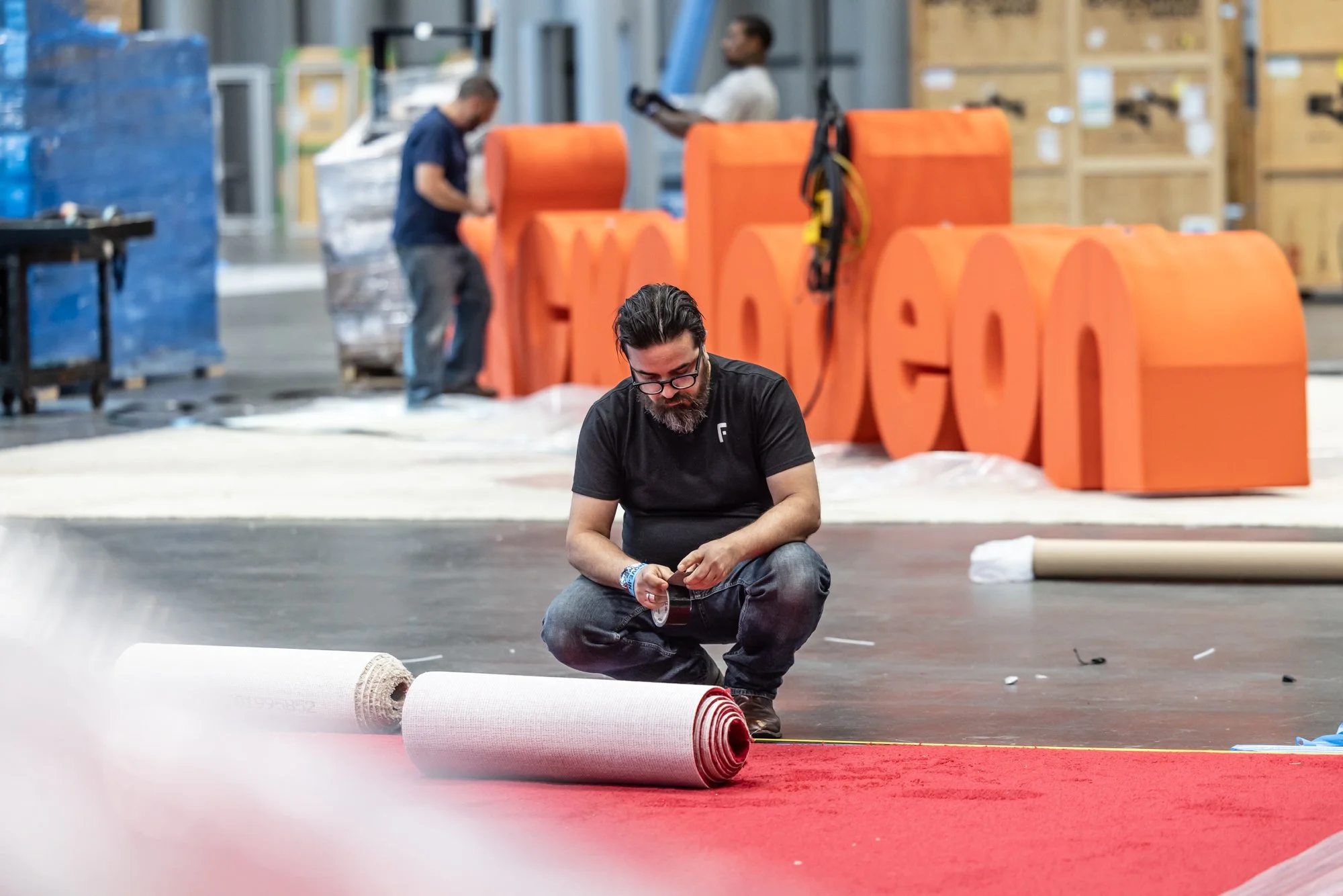 Man rolling out a red carpet in a warehouse event setup with large orange props and workers in the background
