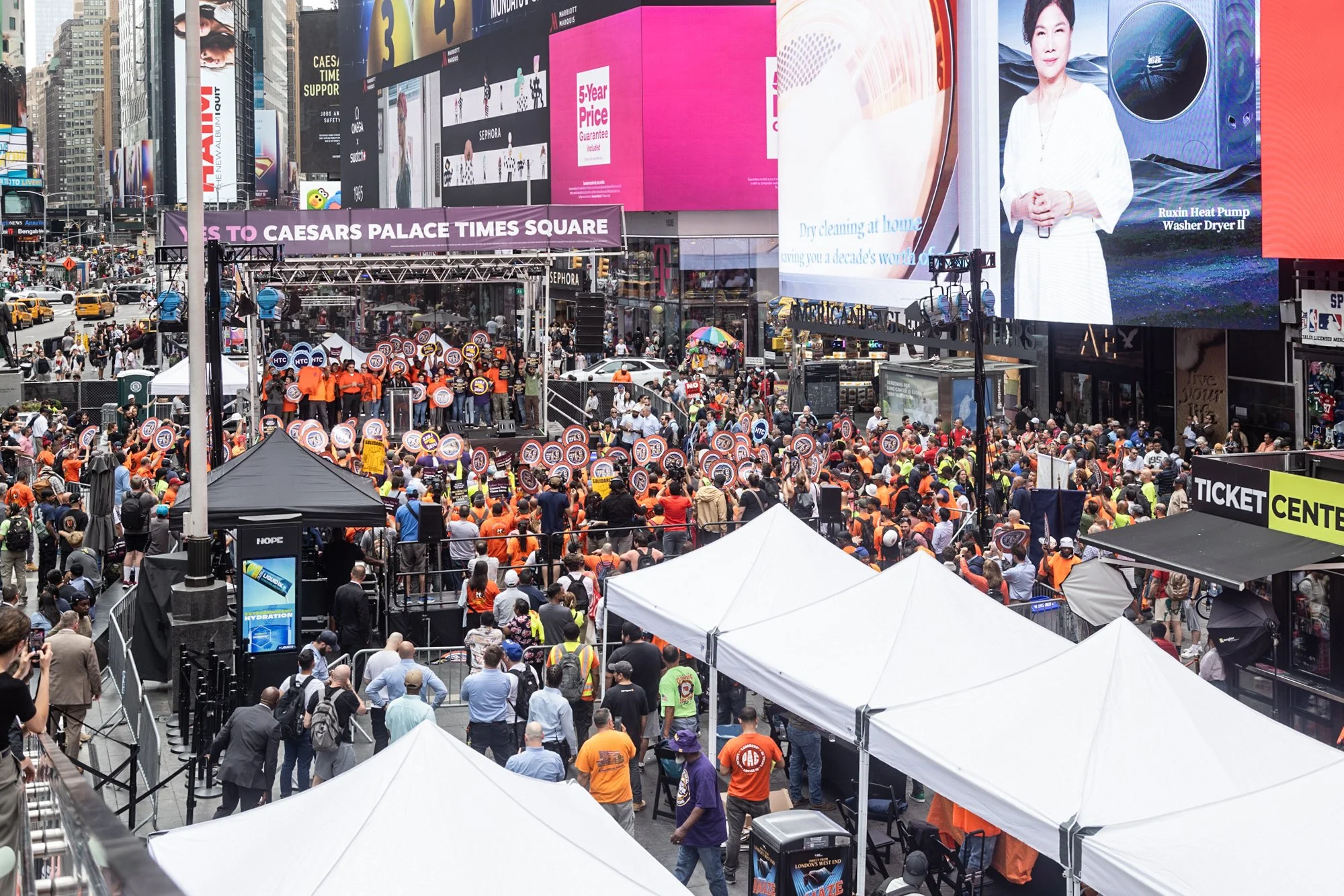 Crowd gathered in Times Square, New York City, with numerous electronic billboards displaying advertisements and signs including a banner that reads 'STEPS TO CAESARS PALACE TIMES SQUARE.' People are walking and standing around, some wearing orange s