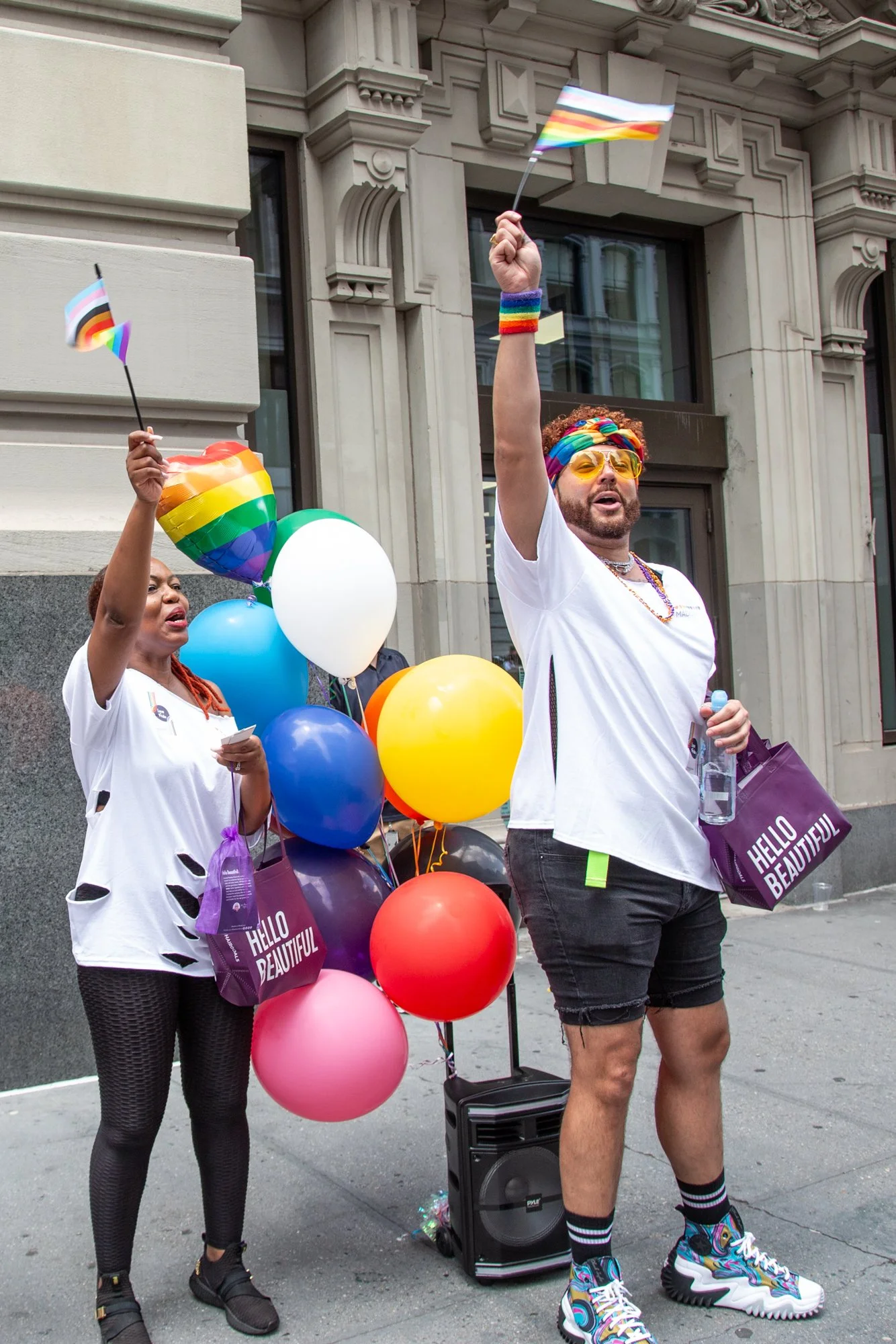 Two people celebrate Pride with rainbow flags, colorful balloons, and rainbow-themed clothing outside a building.