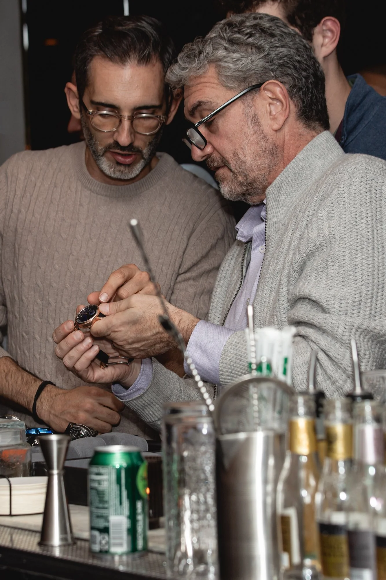 Two men are closely examining a wristwatch together in a setting that appears to be a workshop or laboratory, with various tools and equipment on the table in front of them.