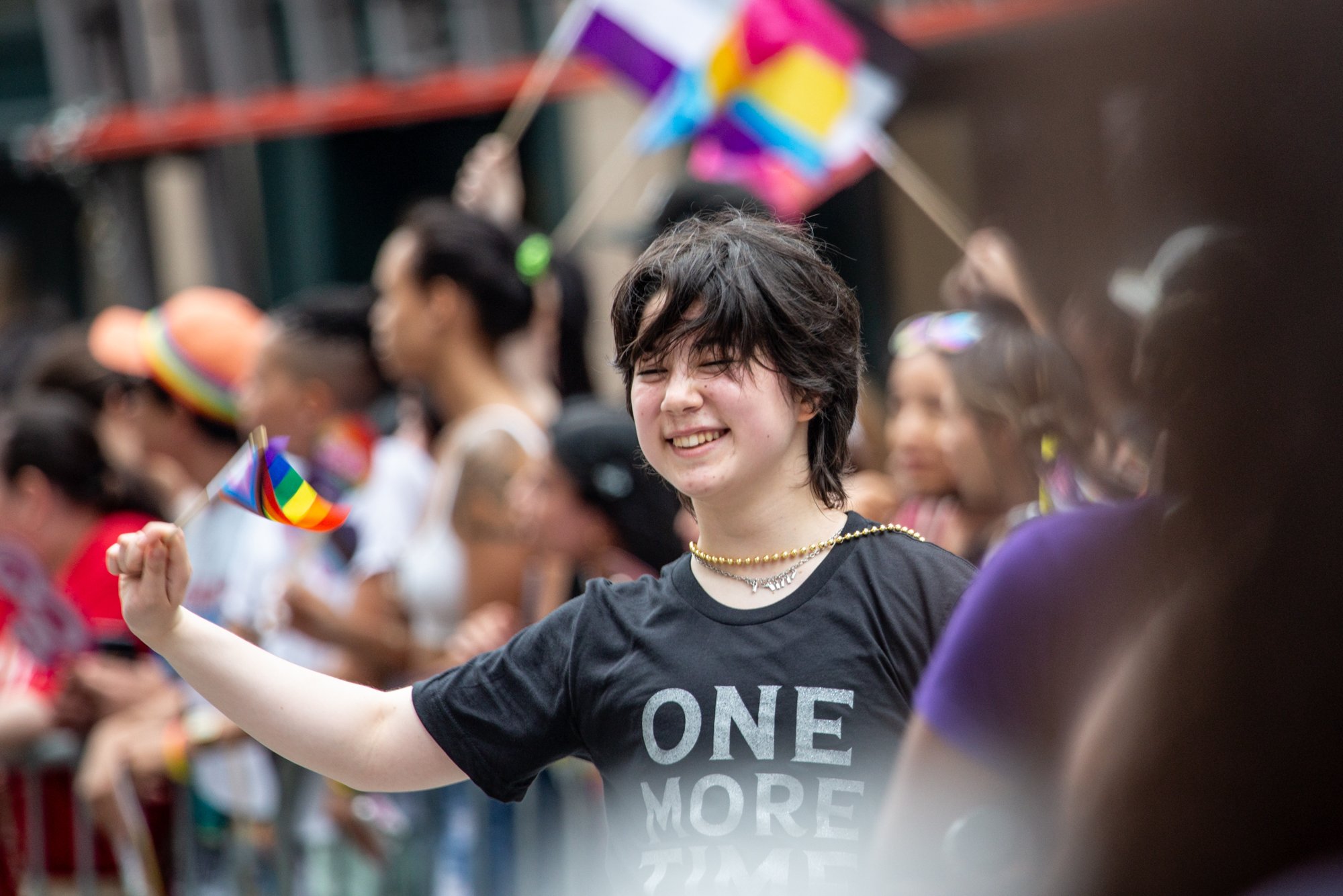 Smiling person in a black “ONE MORE” shirt waving a rainbow Pride flag in a crowd.