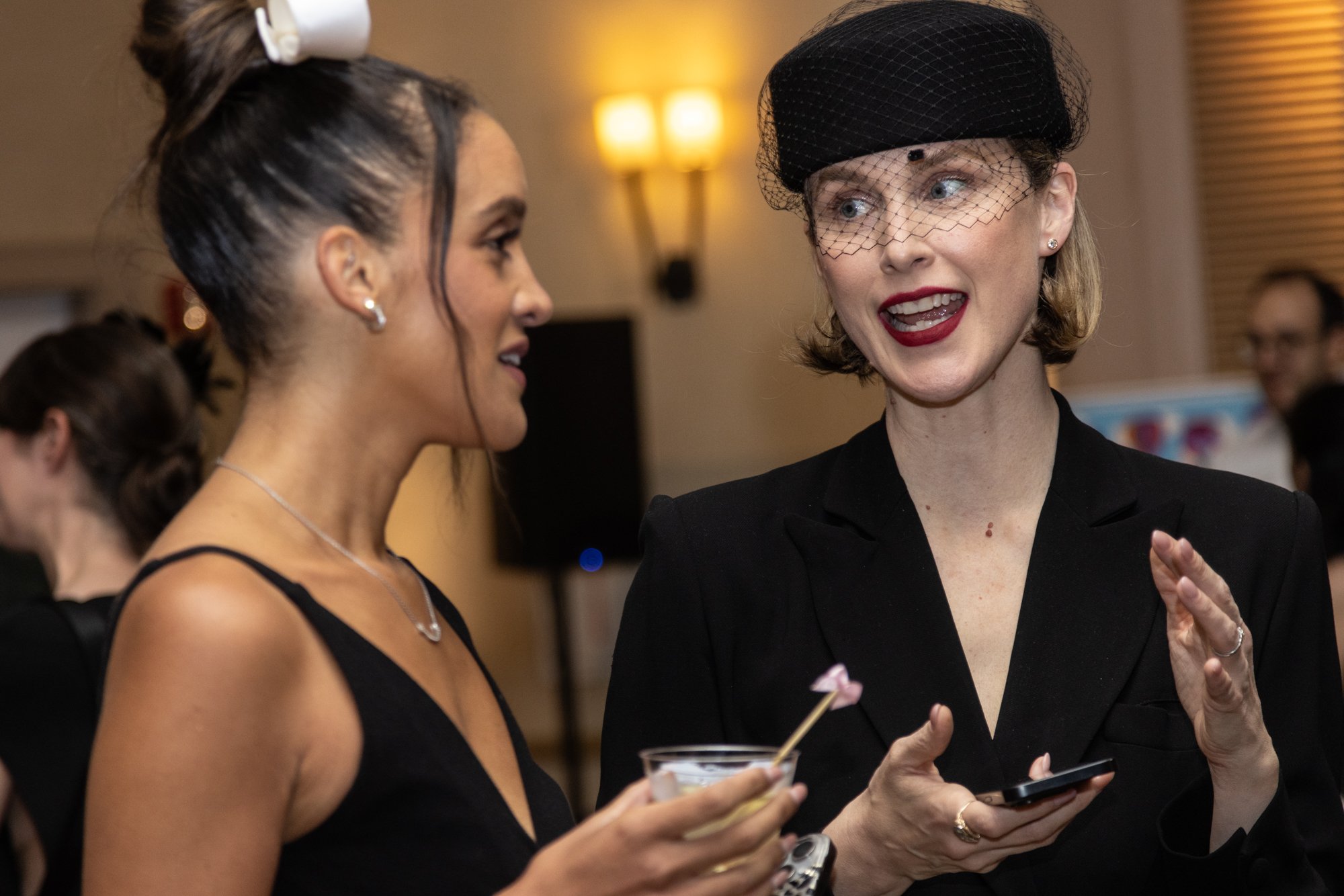 Two women engaged in conversation at an indoor social event. The woman on the left has dark hair in an updo, wearing a black dress, and pearl earrings. The woman on the right has short hair, wearing a black outfit with a netted black hat, bright red 