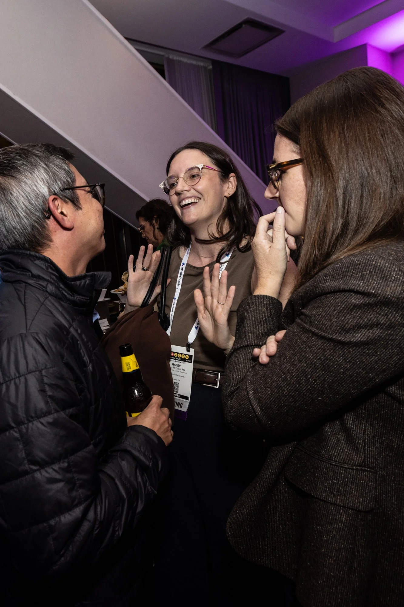 Three women and one man engaged in conversation at an indoor event, with one woman wearing glasses and a conference badge, holding a beverage, in a dimly lit room with purple accent lighting.