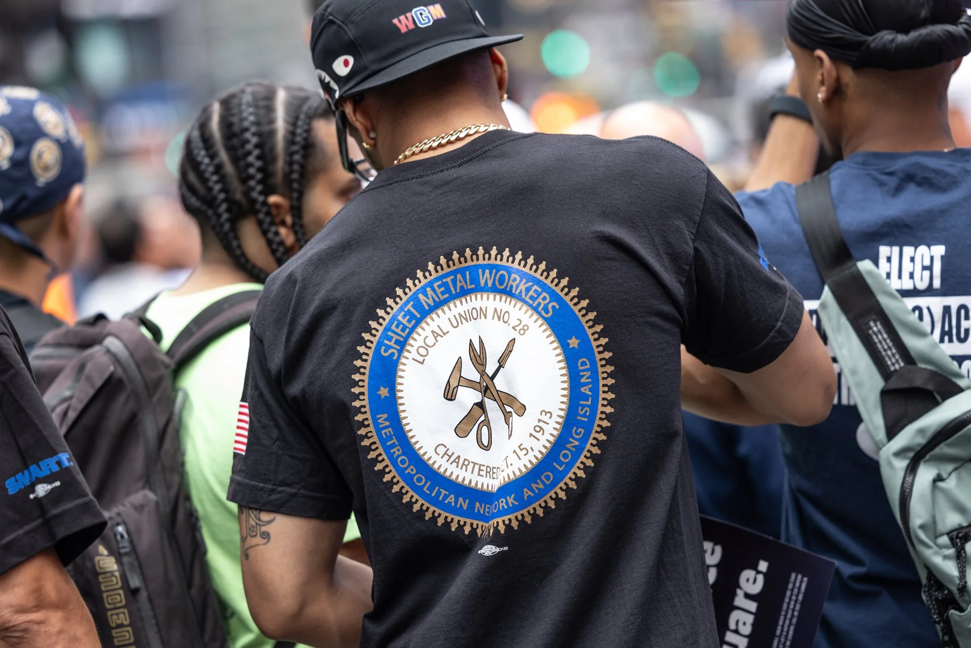 People gathered outdoors, one person wearing a black shirt with a logo for Sheet Metal Workers Local Union No. 28, and others with backpacks and head coverings, likely participating in a protest or rally.