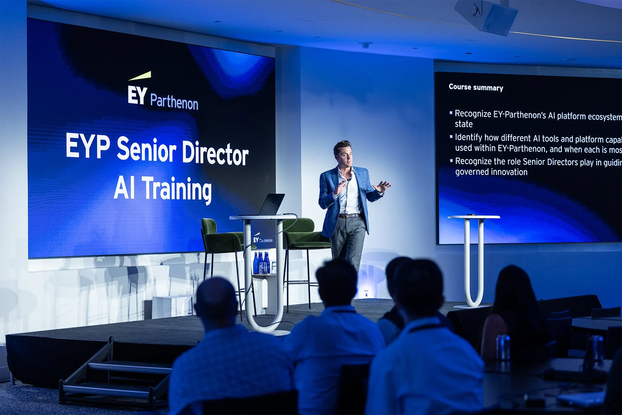 A man in a blue blazer and white shirt giving a presentation at a conference, standing between two screens displaying a course titled 'EYP Senior Director AI Training' and course details, with audience members visible in the foreground.