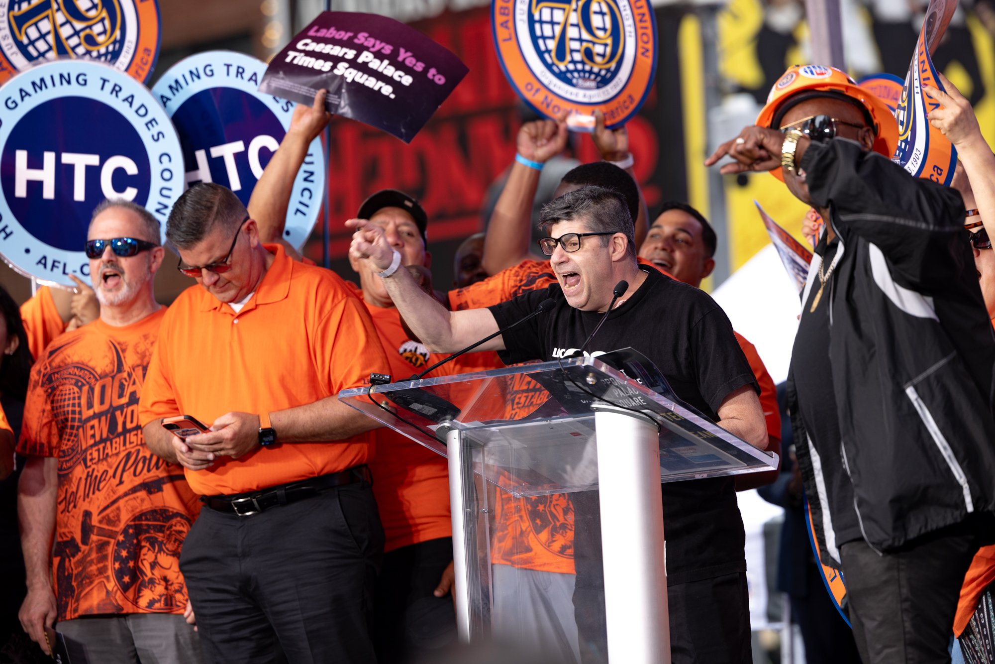 People gathered at a rally, some wearing orange shirts, with banners and signs supporting the Teamsters union, including HTC logos and a sign saying "Labor Says Yes to Caesars Palace Times Square." A man with glasses and a black shirt is speaking at 