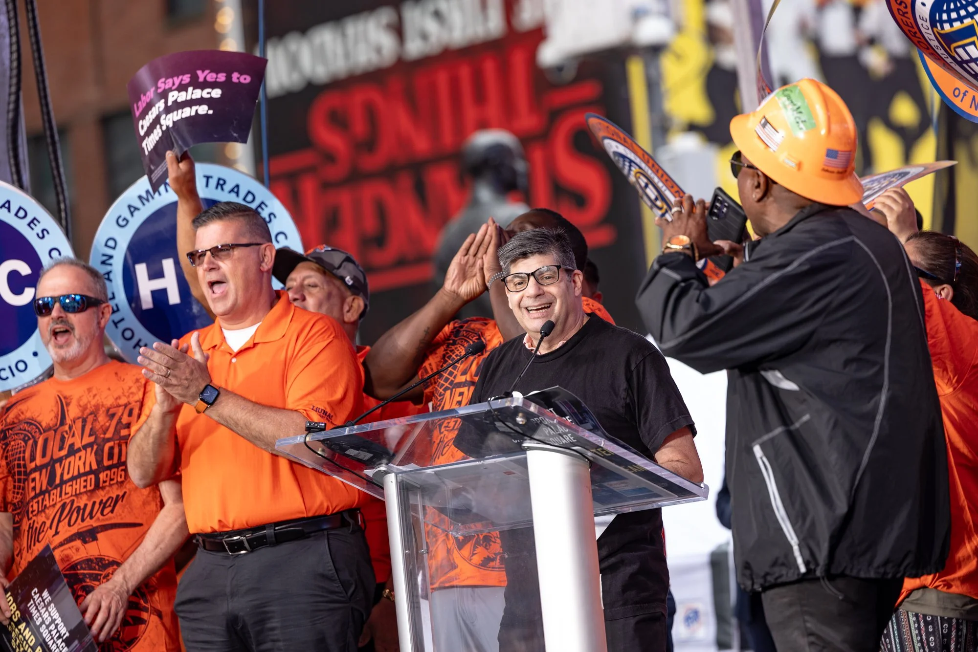 Group of people, including men and women, wearing orange shirts and a black shirt, with some wearing sunglasses and hard hats, gathered on an outdoor stage during a rally or protest, with signs and banners in the background supporting labor and worke