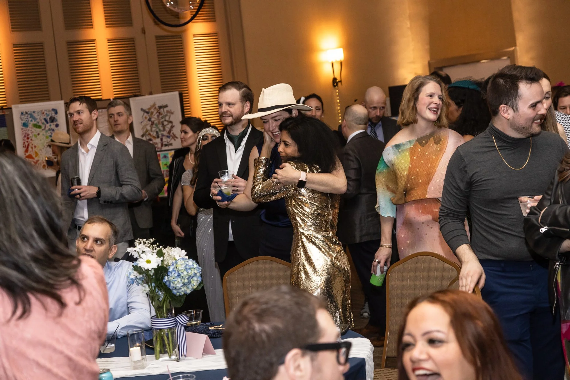 People are gathered in a ballroom during a social event or party, some are standing and mingling while others are seated at tables with floral centerpieces. The decor includes warm lighting and artwork on the walls.