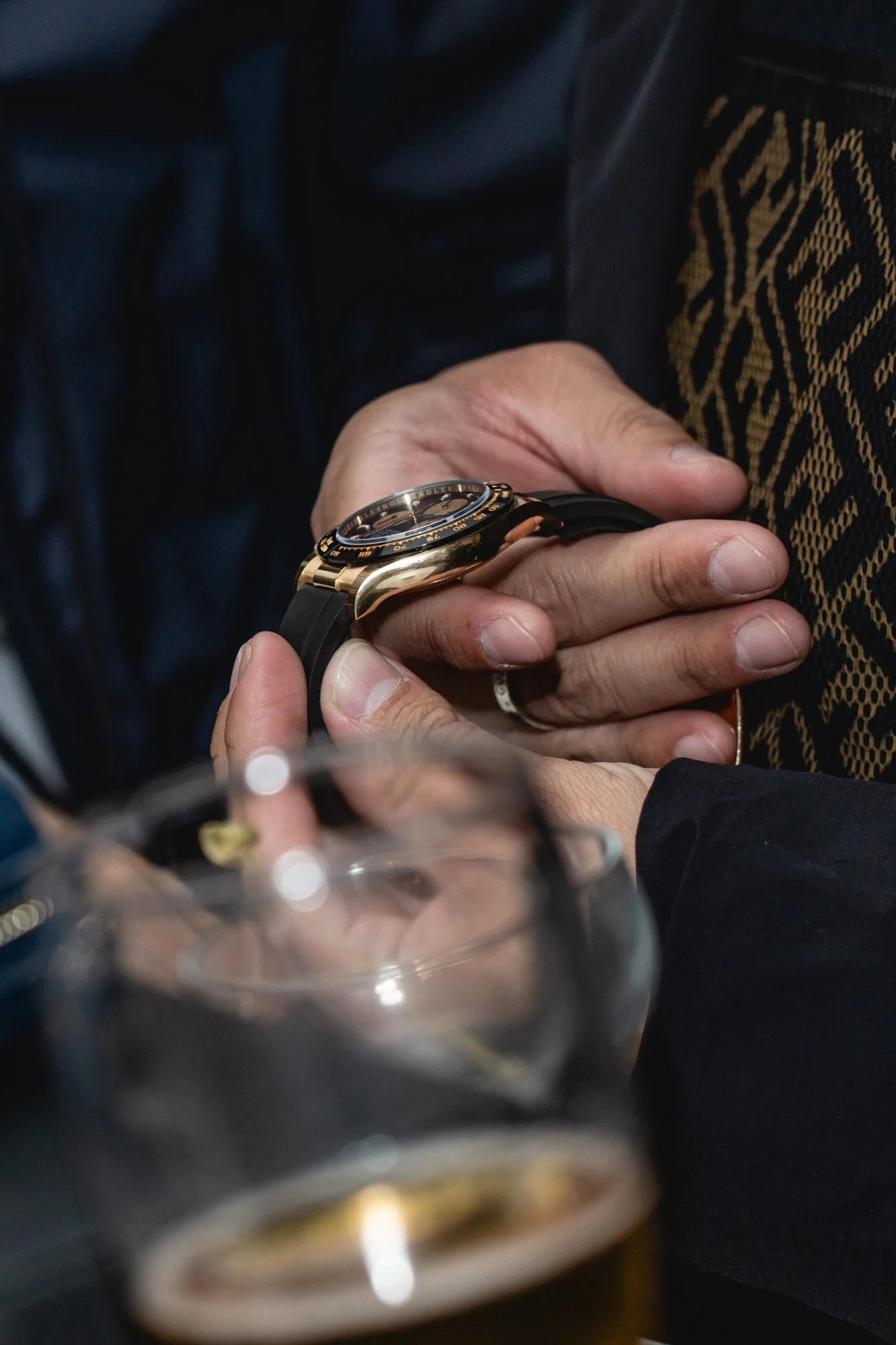 Close-up of a person's hand holding a luxury wristwatch with a black strap and gold accents, with a glass of bubbly in the foreground.