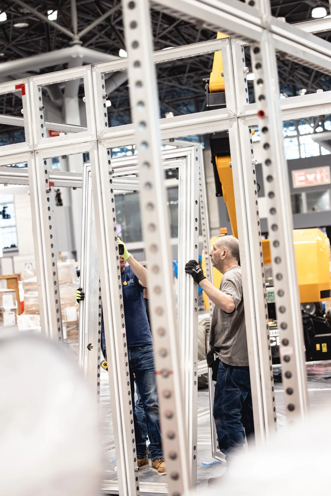 Two workers wearing gloves assemble a white metal industrial framework inside a factory.
