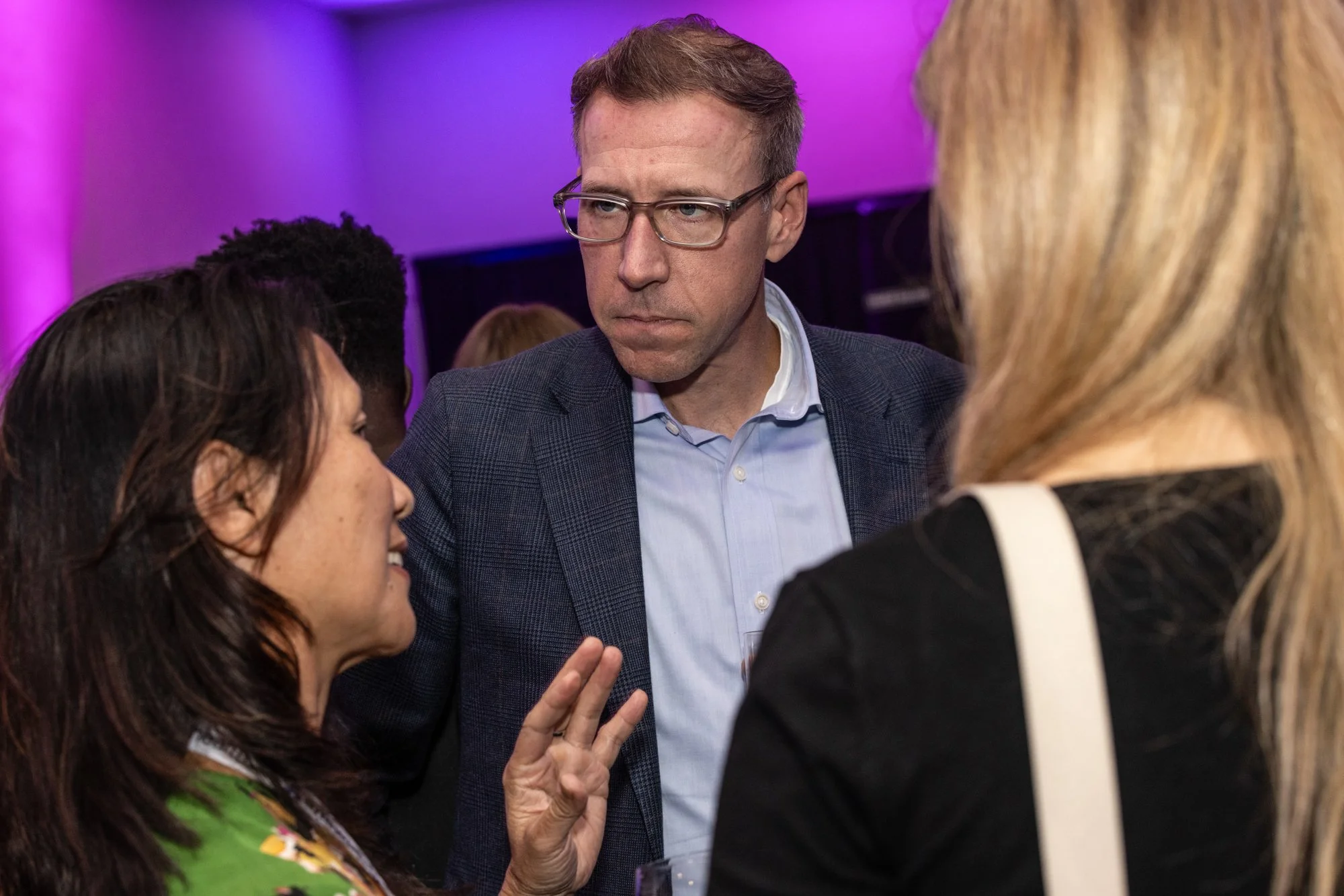 A man with glasses in a suit is looking and listening to a group of women talking in a lively conversation at an indoor event with purple and pink lighting.