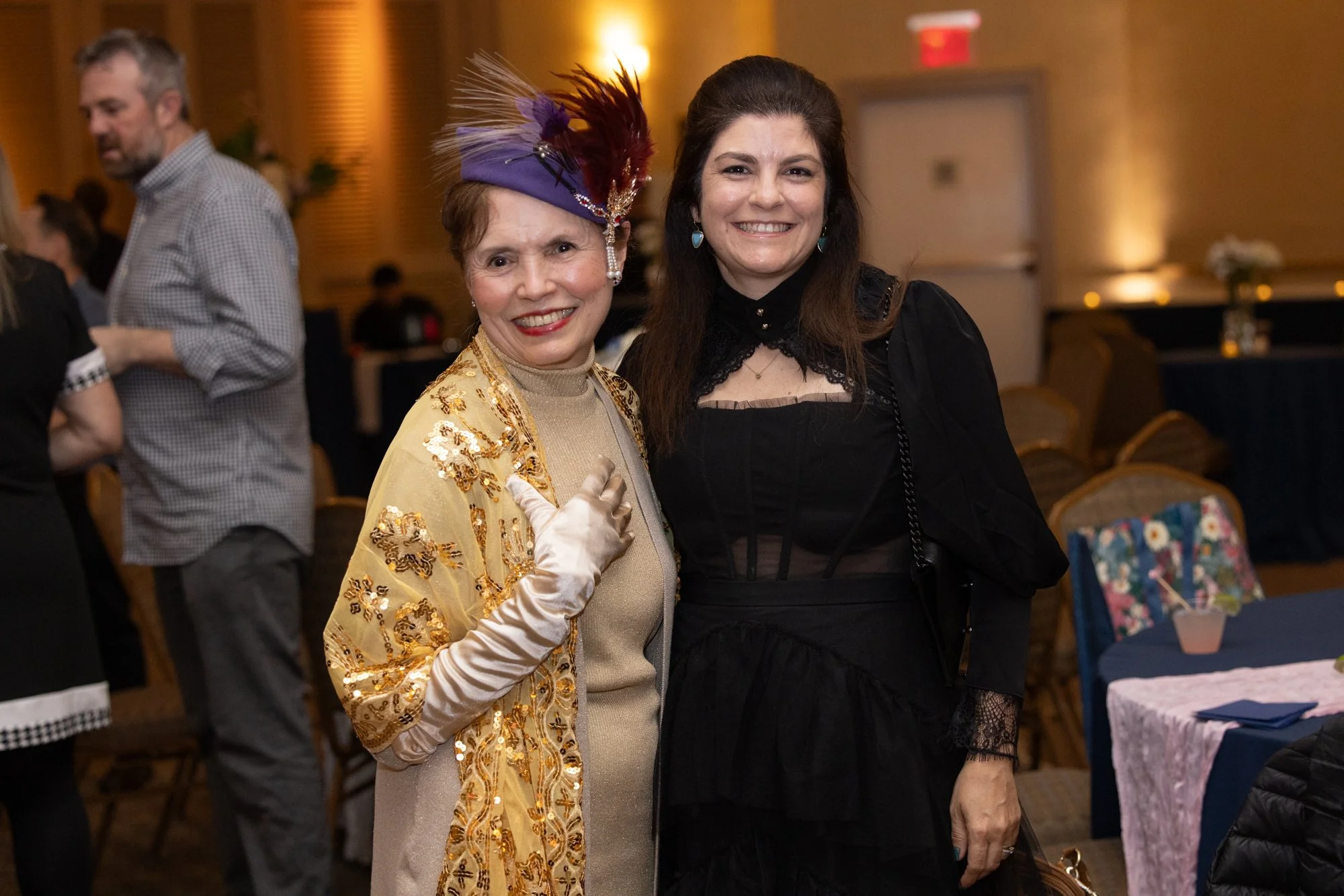 Two women smiling at a social event, one dressed in a gold embellished outfit with a purple hat with feathers, the other in a black lace dress.