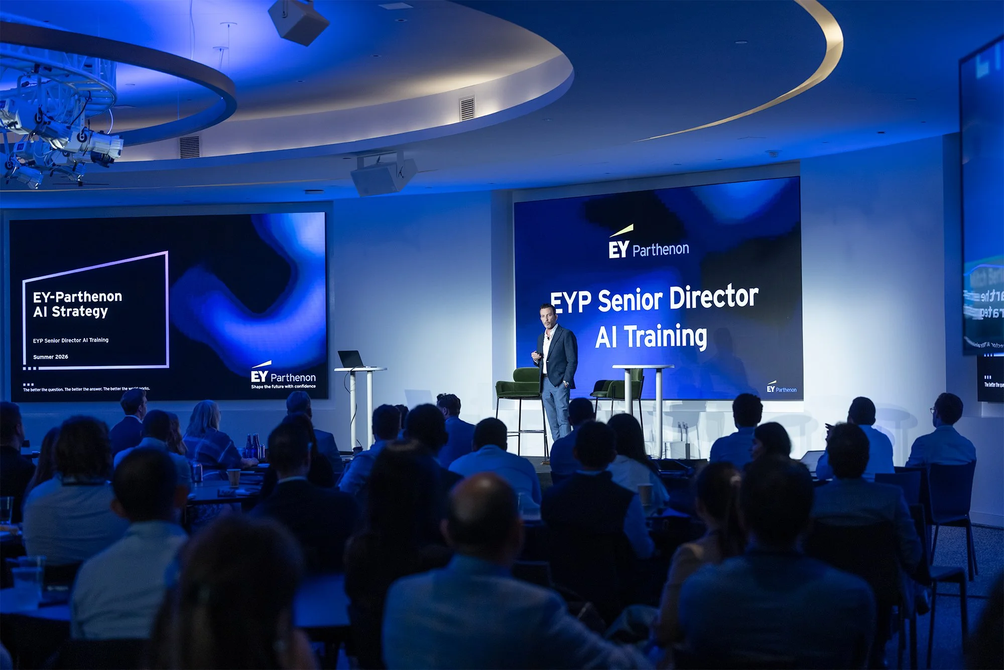 A man in a suit presenting on stage at an event called EY-Parthenon AI Strategy, with audience members seated and listening. The stage has large screens displaying the event title and logo, and the room has a modern design with blue and white lightin