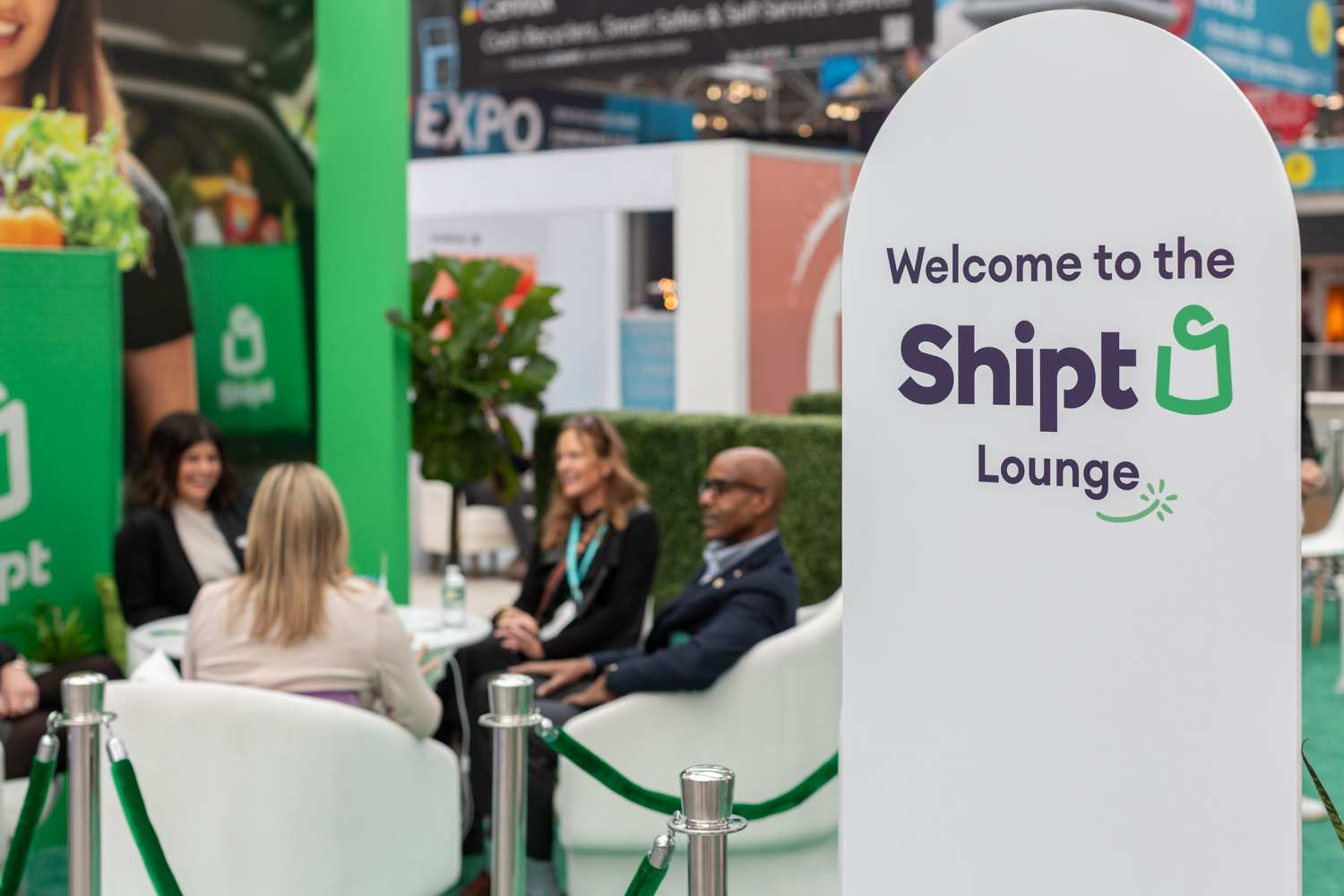People sitting and talking at the Shirt Lounge booth with a welcoming sign in the foreground.