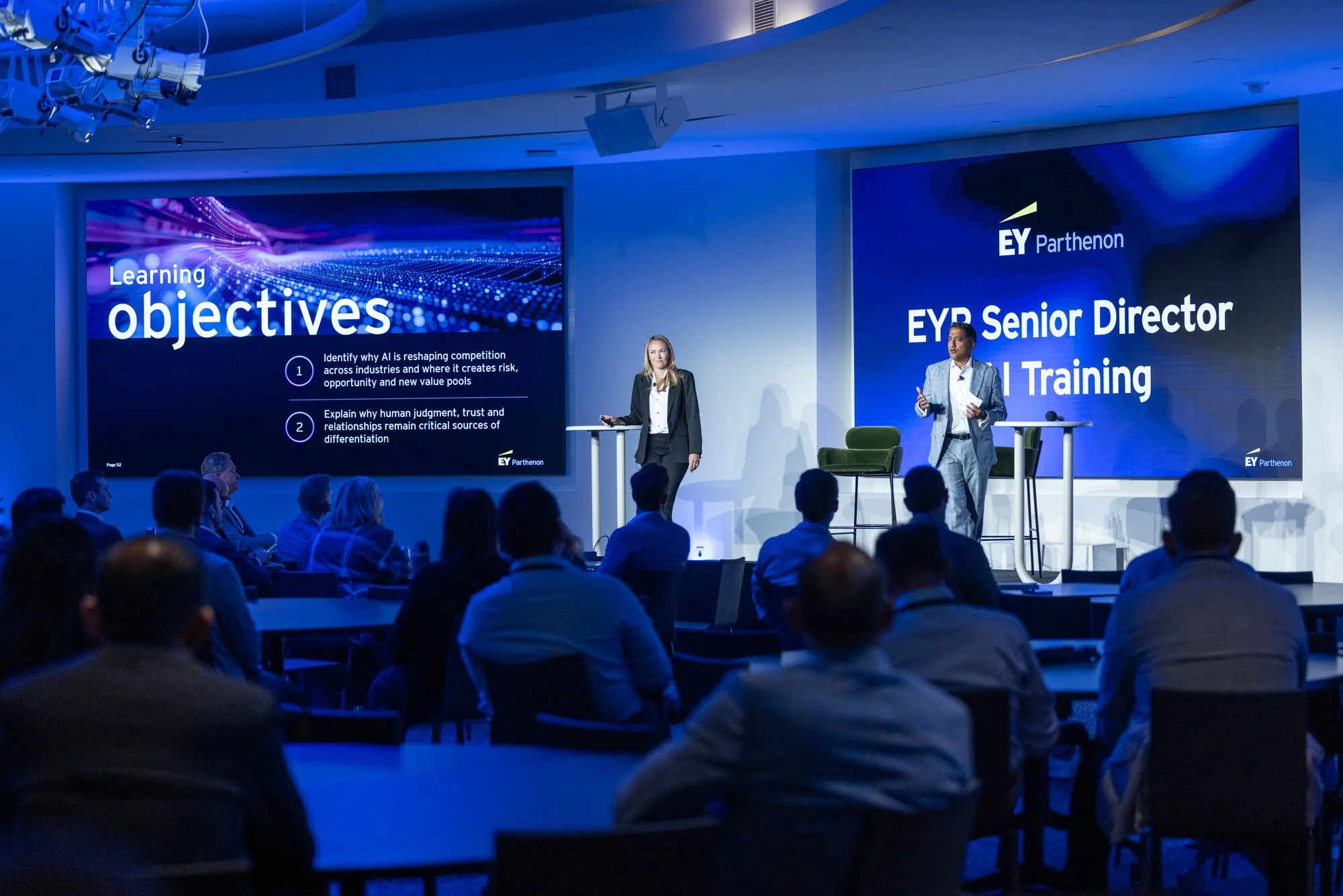 A conference room filled with an audience listening to a presentation. Two speakers stand at the front, one woman and one man, with large digital screens behind them. The screens display information about learning objectives and a company logo, EY Pa