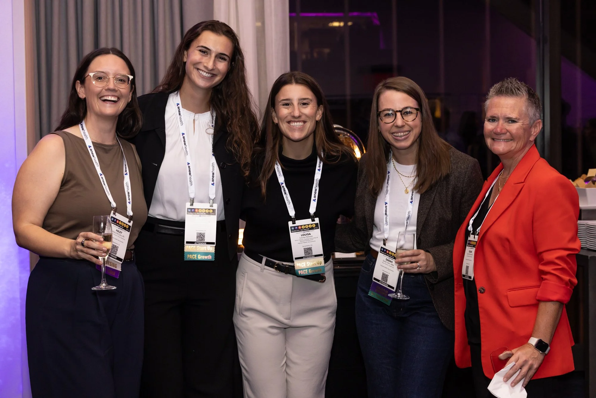 Five women at a conference, wearing name tags and holding glasses of champagne, standing closely together and smiling for the camera in a well-lit indoor space.