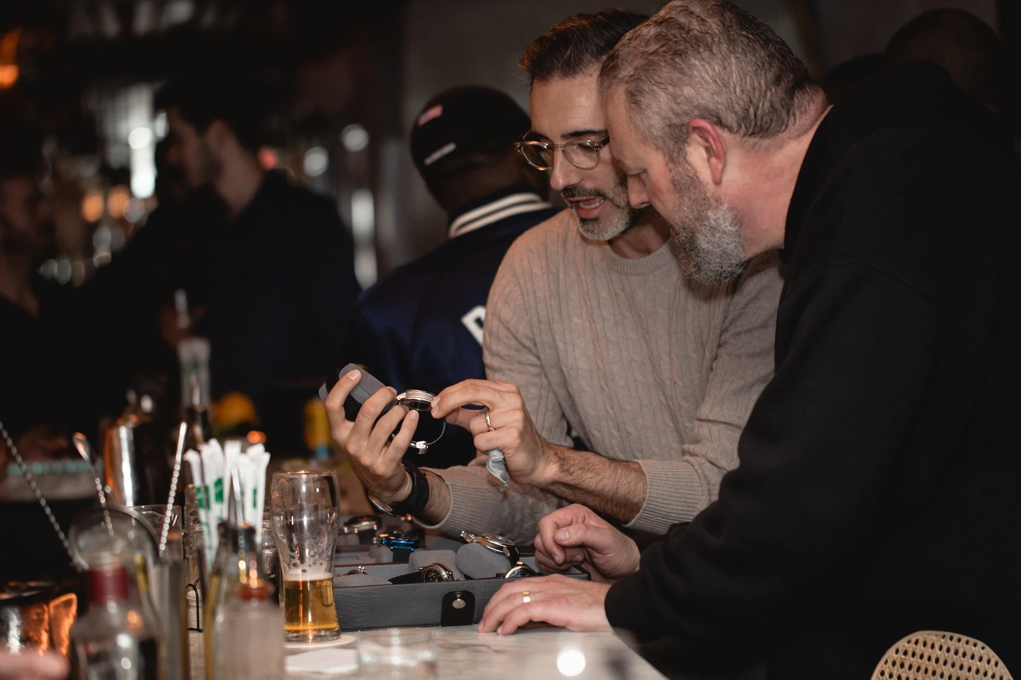 Two men examining a watch at a bar or restaurant, surrounded by glasses and bar items.
