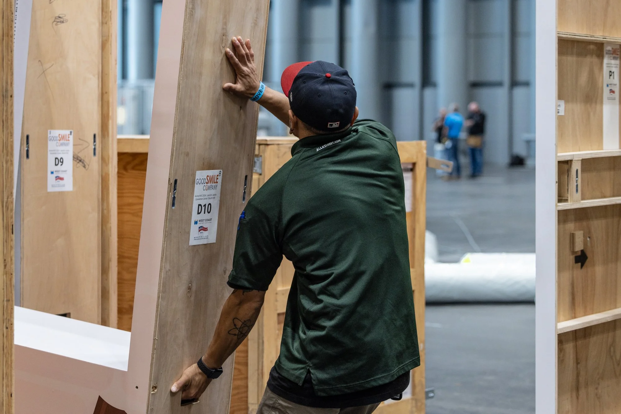Worker in a baseball cap moving a labeled wooden exhibit panel inside a trade show booth.