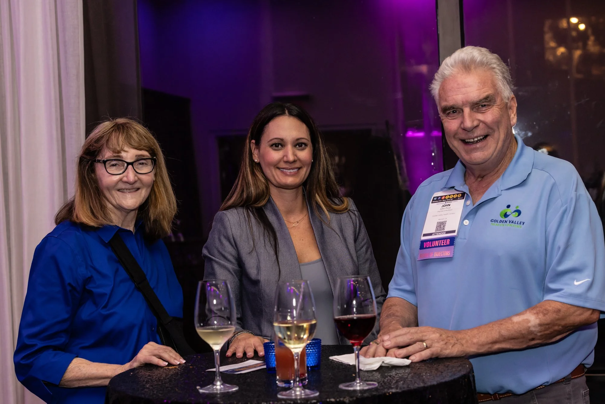 Three people standing together at a social event, with wine glasses on a table in front of them. The woman on the left has shoulder-length red hair, glasses, and is wearing a blue shirt. The woman in the middle has long brown hair, and is wearing a g