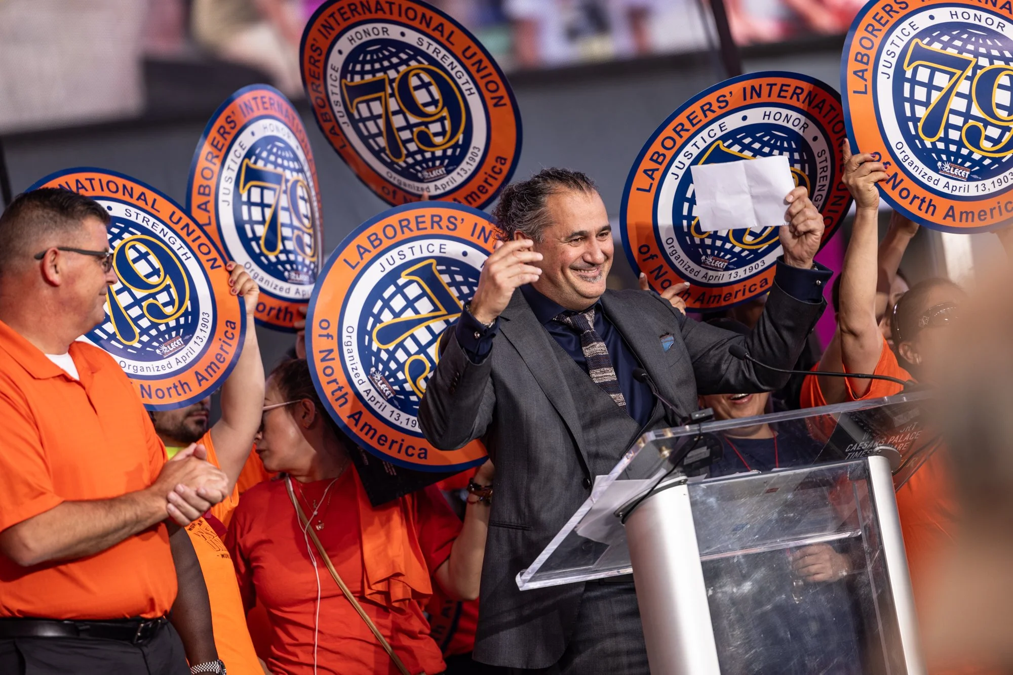A group of people participating in a protest or rally, holding signs that say 'Labourers' International Union of North America' with a logo. One man in a suit is smiling and speaking at a podium, while others are standing nearby.