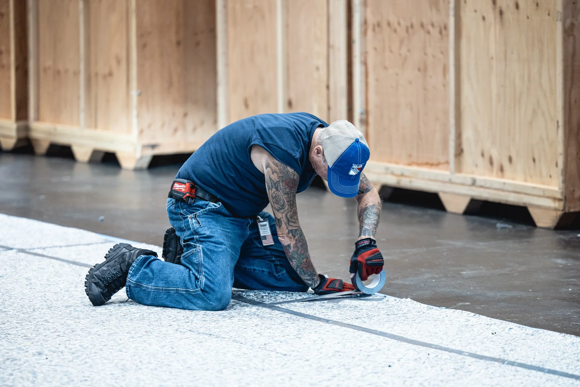 Tattooed construction worker kneeling while applying tape to a floor inside a plywood-framed building