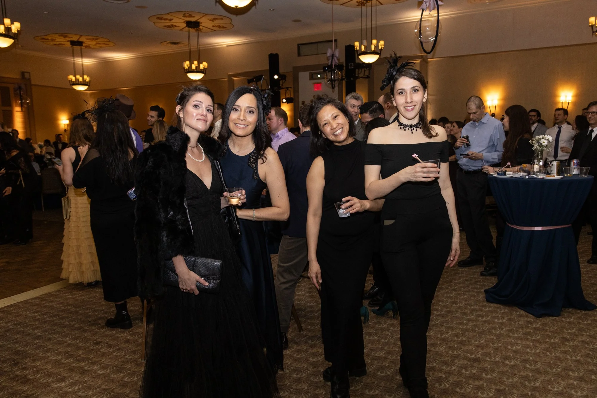 Four women in evening attire standing together at a formal event, smiling at the camera, with a crowd of people in the background in a decorated banquet hall.