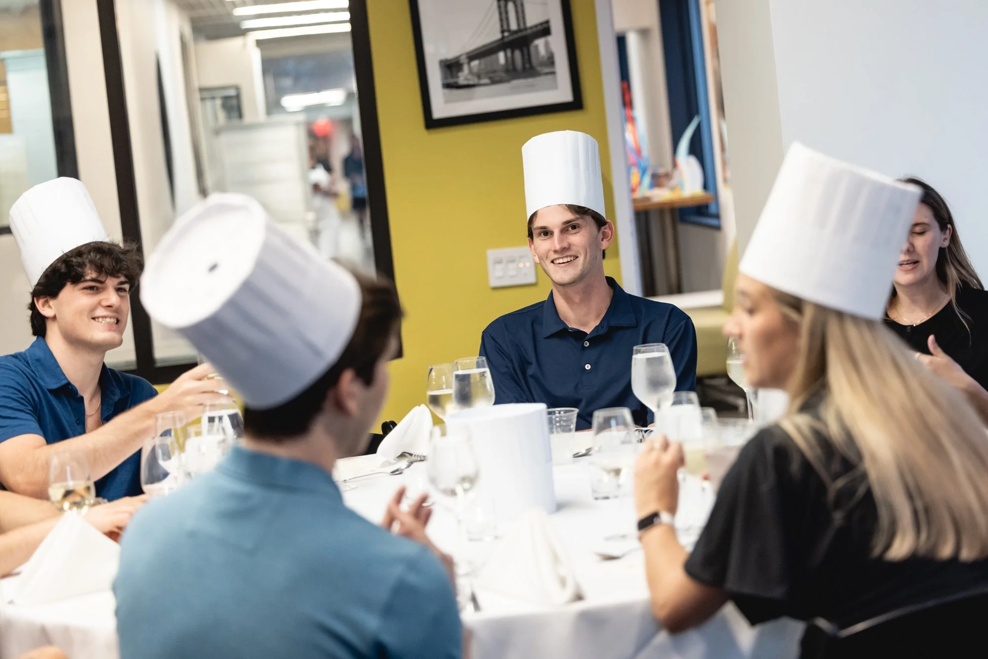 A group of people wearing chef hats dining together at a restaurant table, smiling and engaging in conversation.